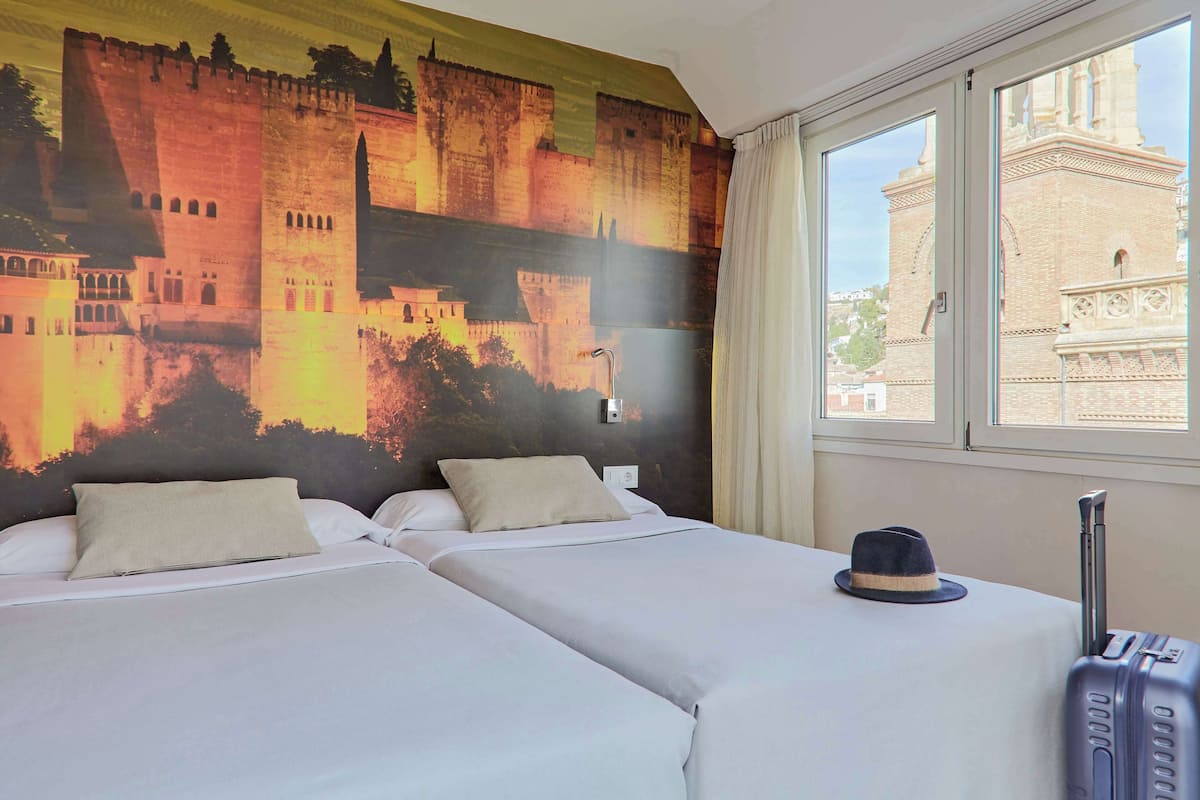 Ett hotellrum med två sängar, en stor väggmålning som föreställer ett slott, ett fönster med utsikt över en byggnad och en resväska.