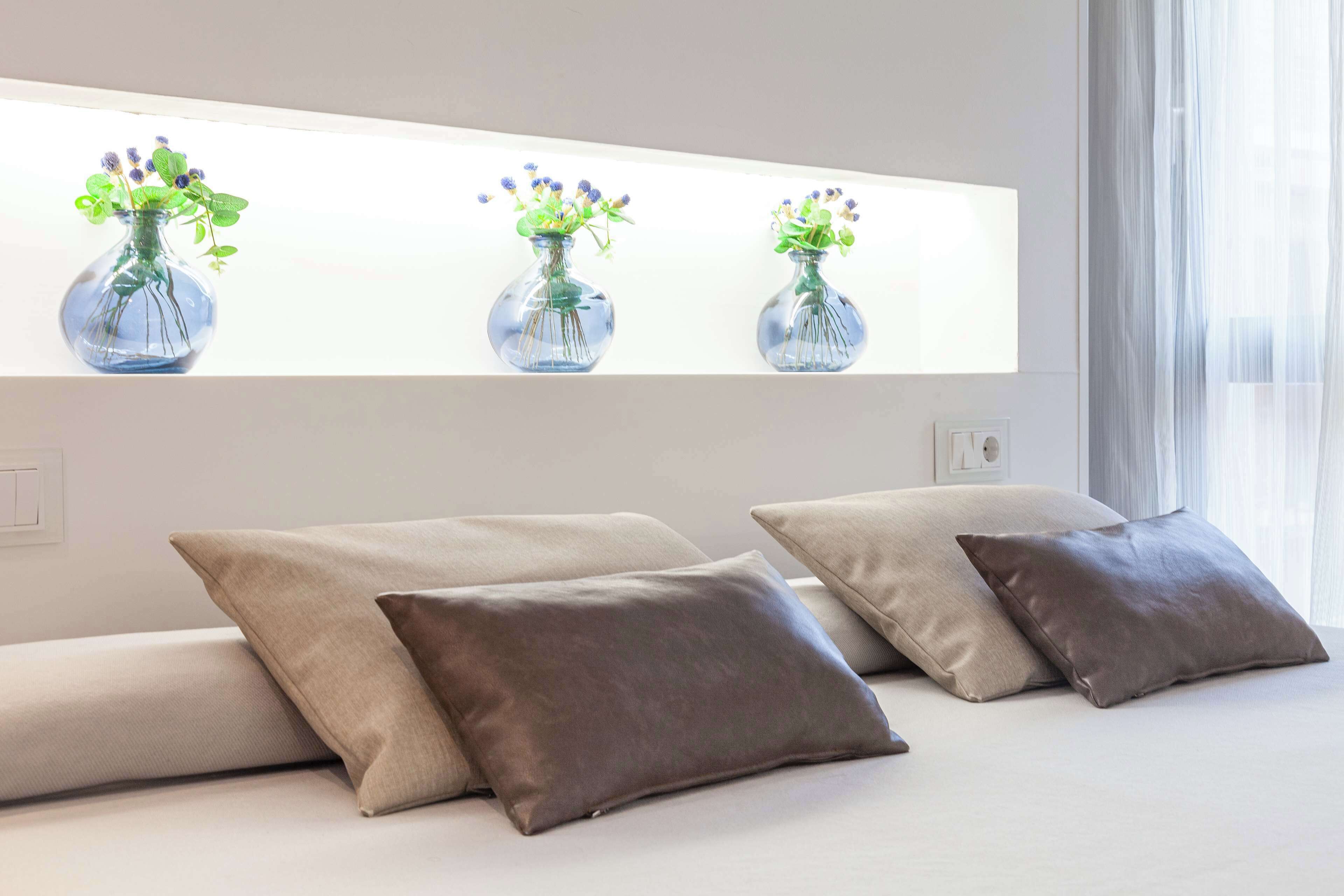 A modern bedroom with a bed, three pillows, and three illuminated vases with flowers on a wall.