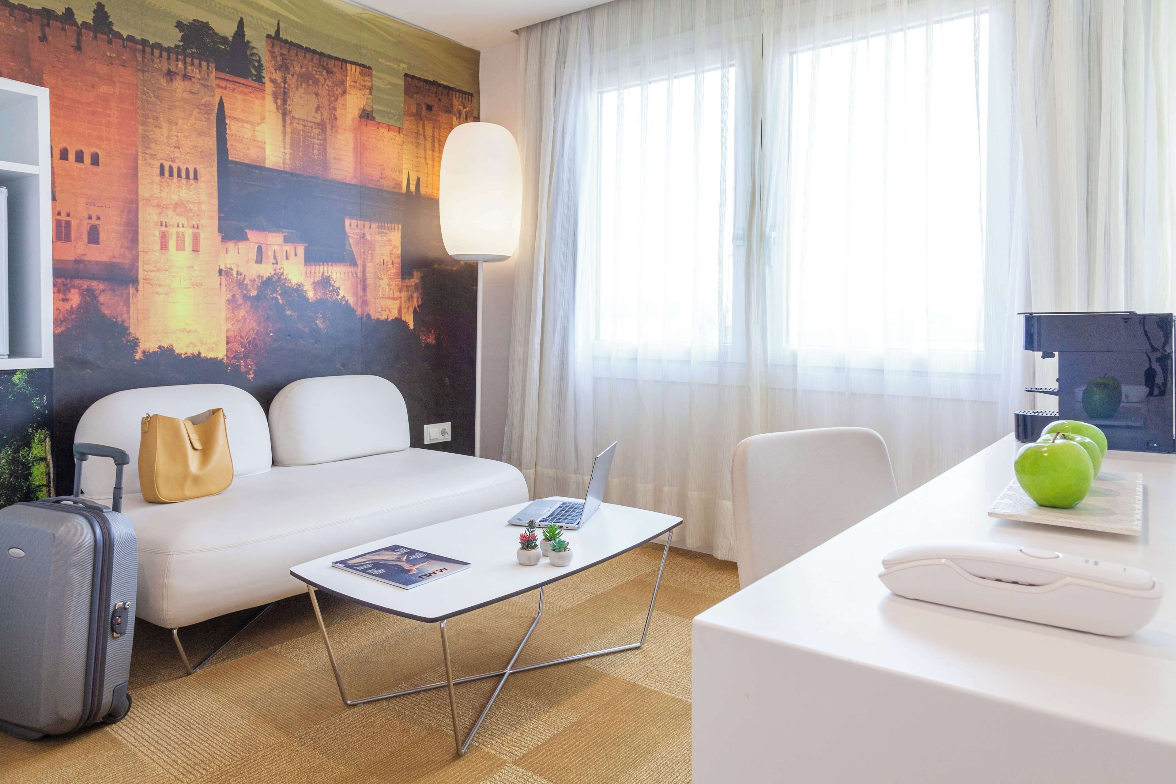 Ein Hotelzimmer mit einem Bett, einem Schreibtisch mit Laptop, einem kleinen Tisch mit Pflanze, einem Koffer und einem Wandbild.