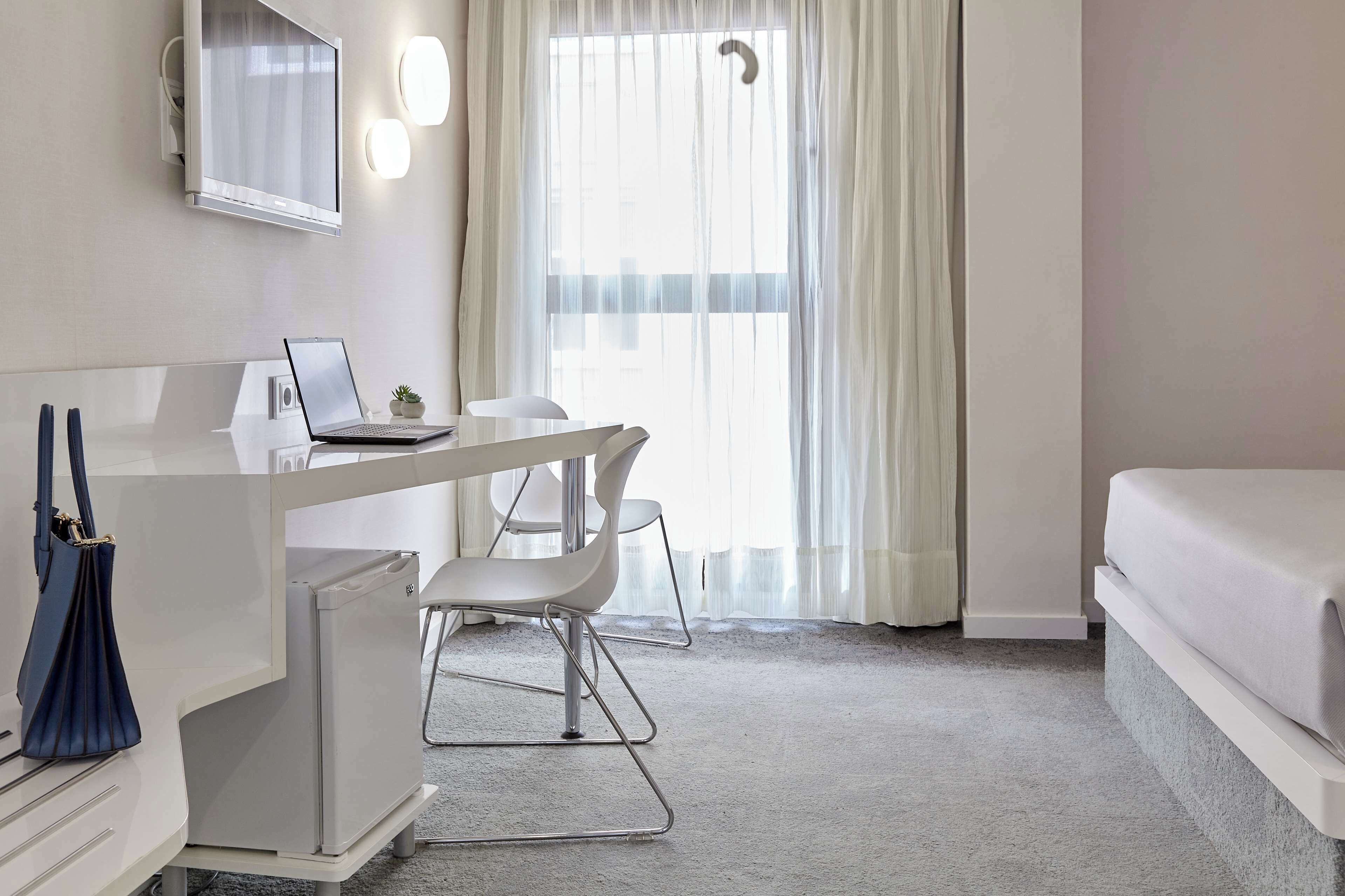 Ein Hotelzimmer mit Schreibtisch, Stuhl, Bett und einem großen Fenster mit durchsichtigen Vorhängen.