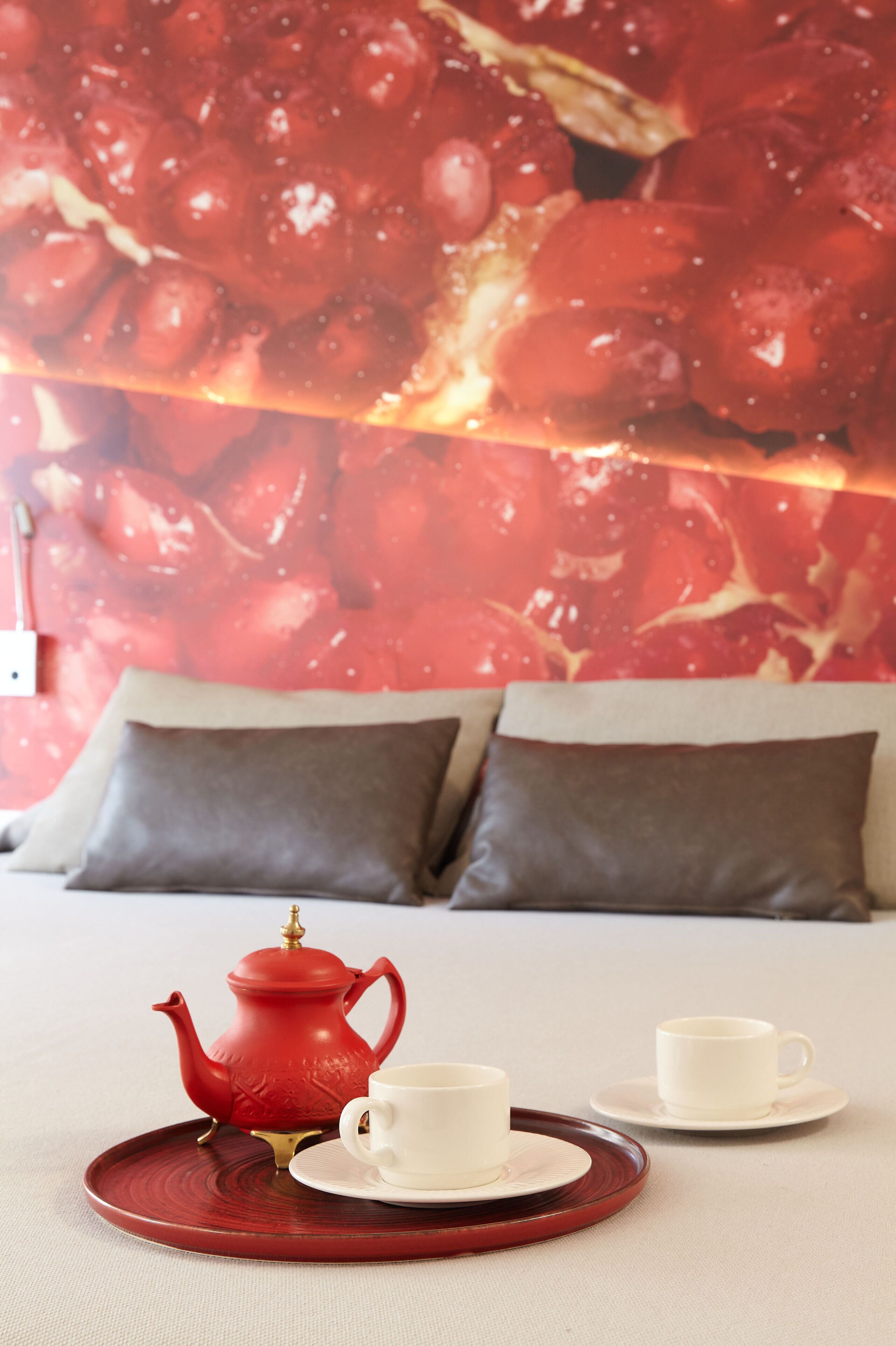 Une chambre à coucher avec un lit, des oreillers, une théière rouge et deux tasses blanches sur un plateau.