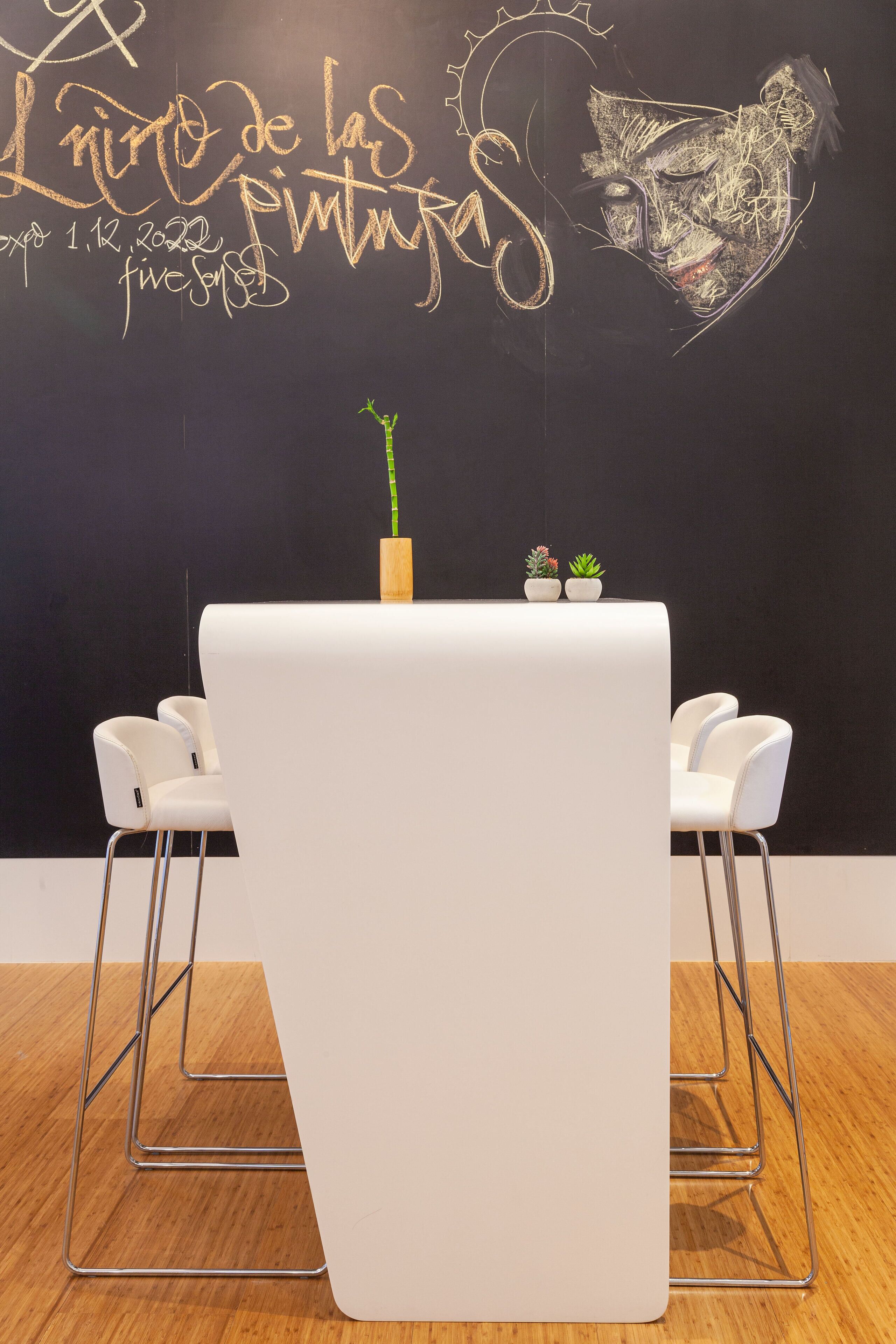 Una habitación moderna con una pizarra que presenta arte y texto hechos con tiza, una mesa blanca con dos plantas en macetas y sillas blancas con estructura metálica.