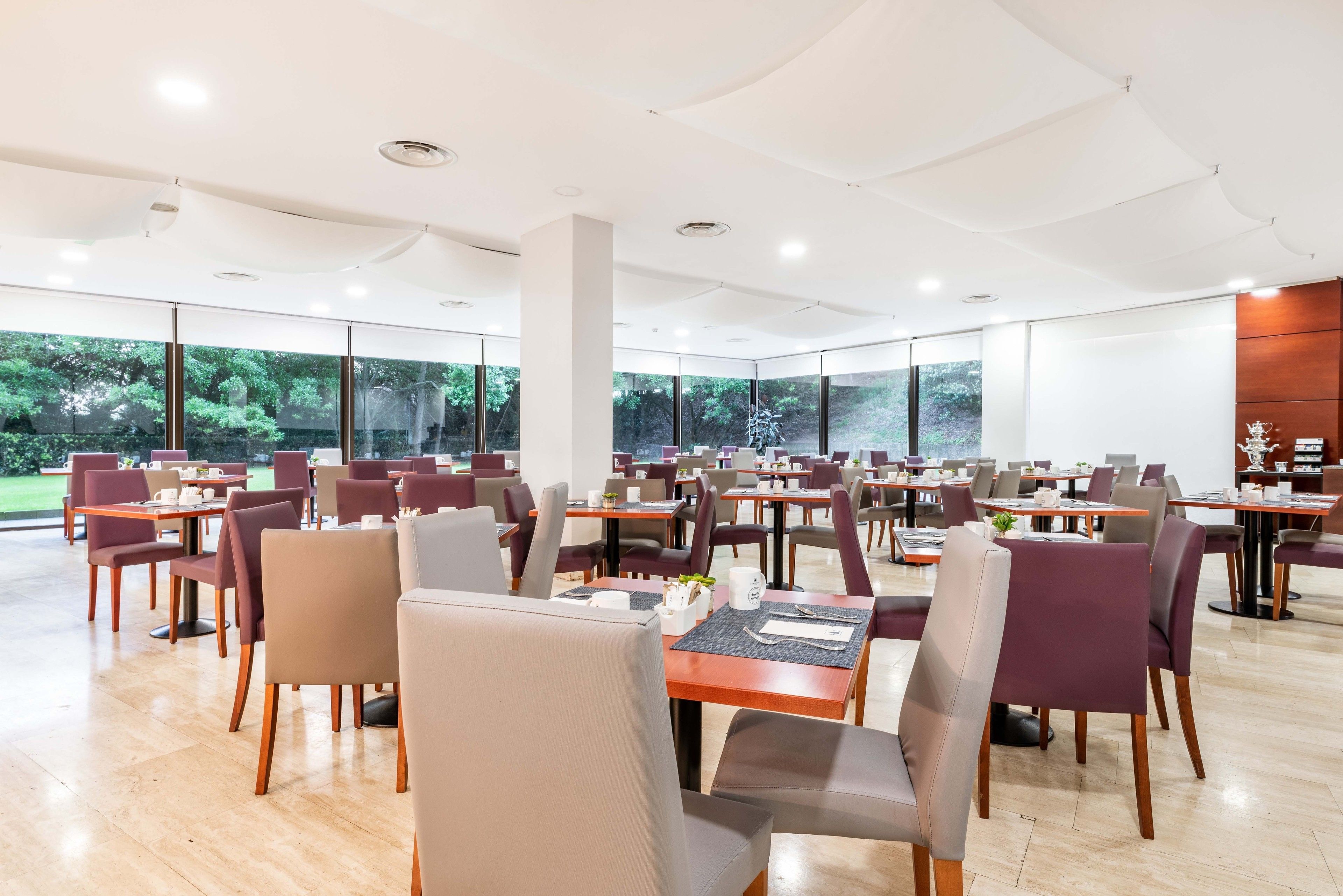 A spacious dining area with multiple tables and chairs, large windows offering a view of greenery, and a modern ceiling design.