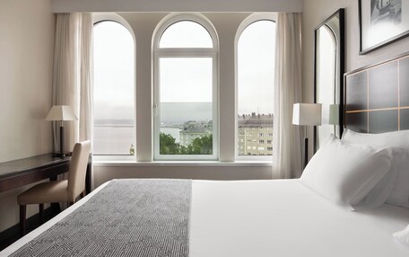 A hotel room with a large bed, a desk, and a chair. There are three windows with views of buildings and a body of water.