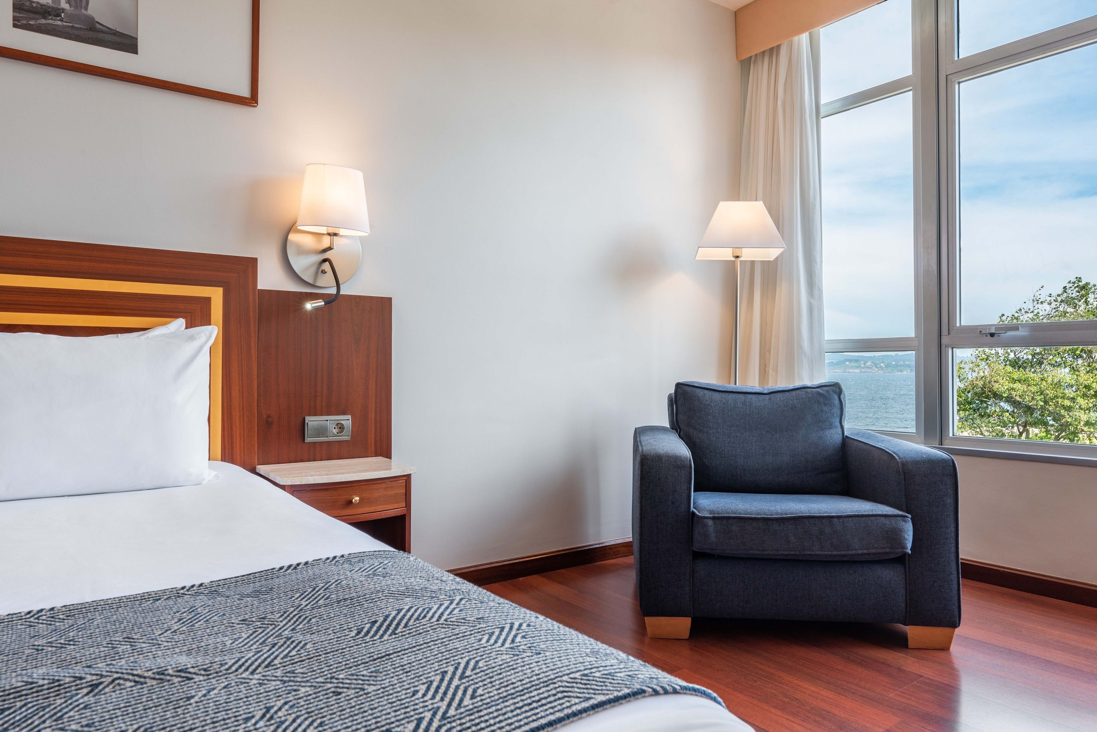 A hotel room with a bed, a nightstand, a lamp, a chair, and a large window with a view.