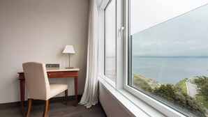 Habitació d'hotel amb escriptori, cadira, làmpada i una finestra gran que ofereix vistes al mar i a la costa.