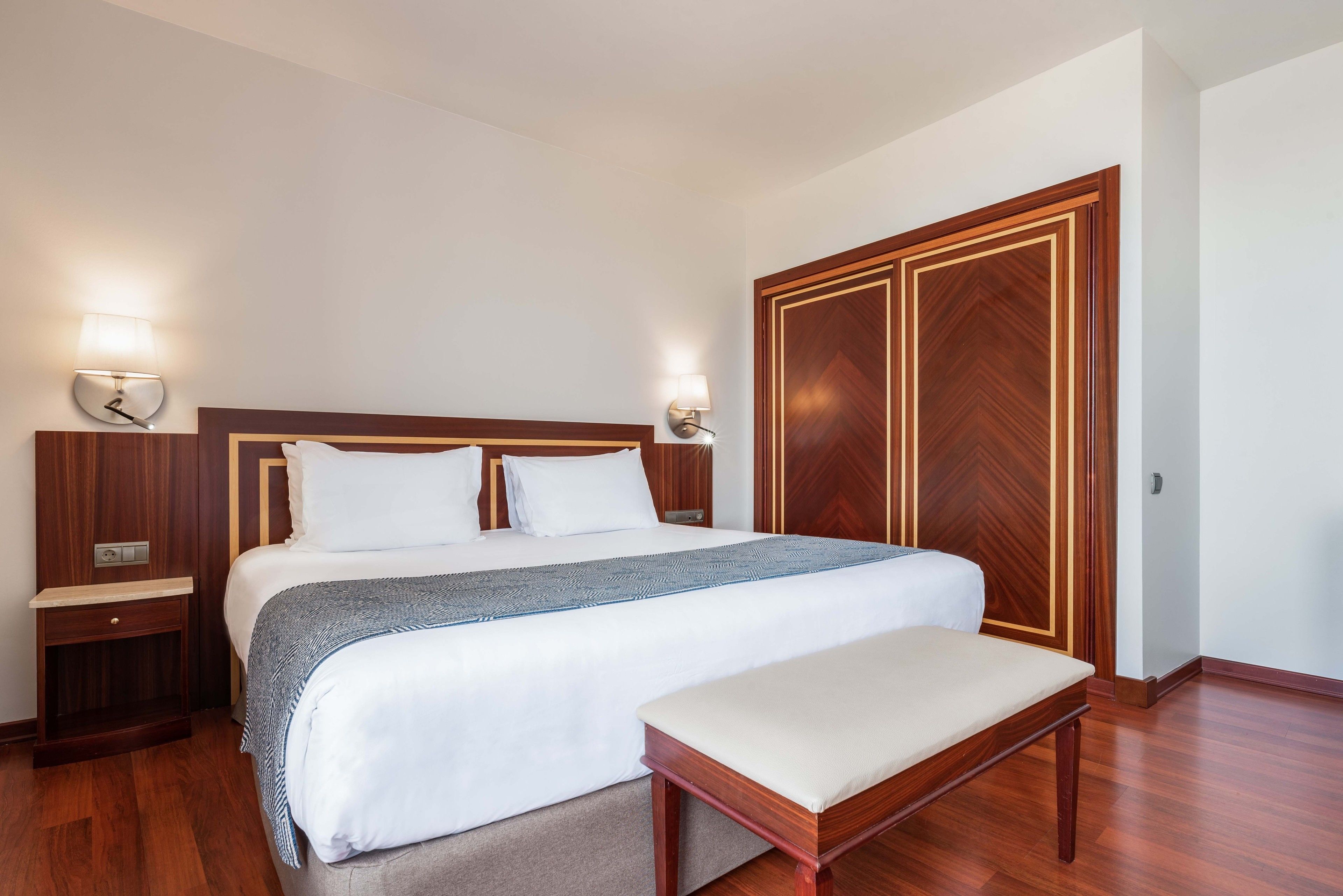 A neatly arranged hotel room with a large bed, bedside tables, a bench, and a wardrobe.