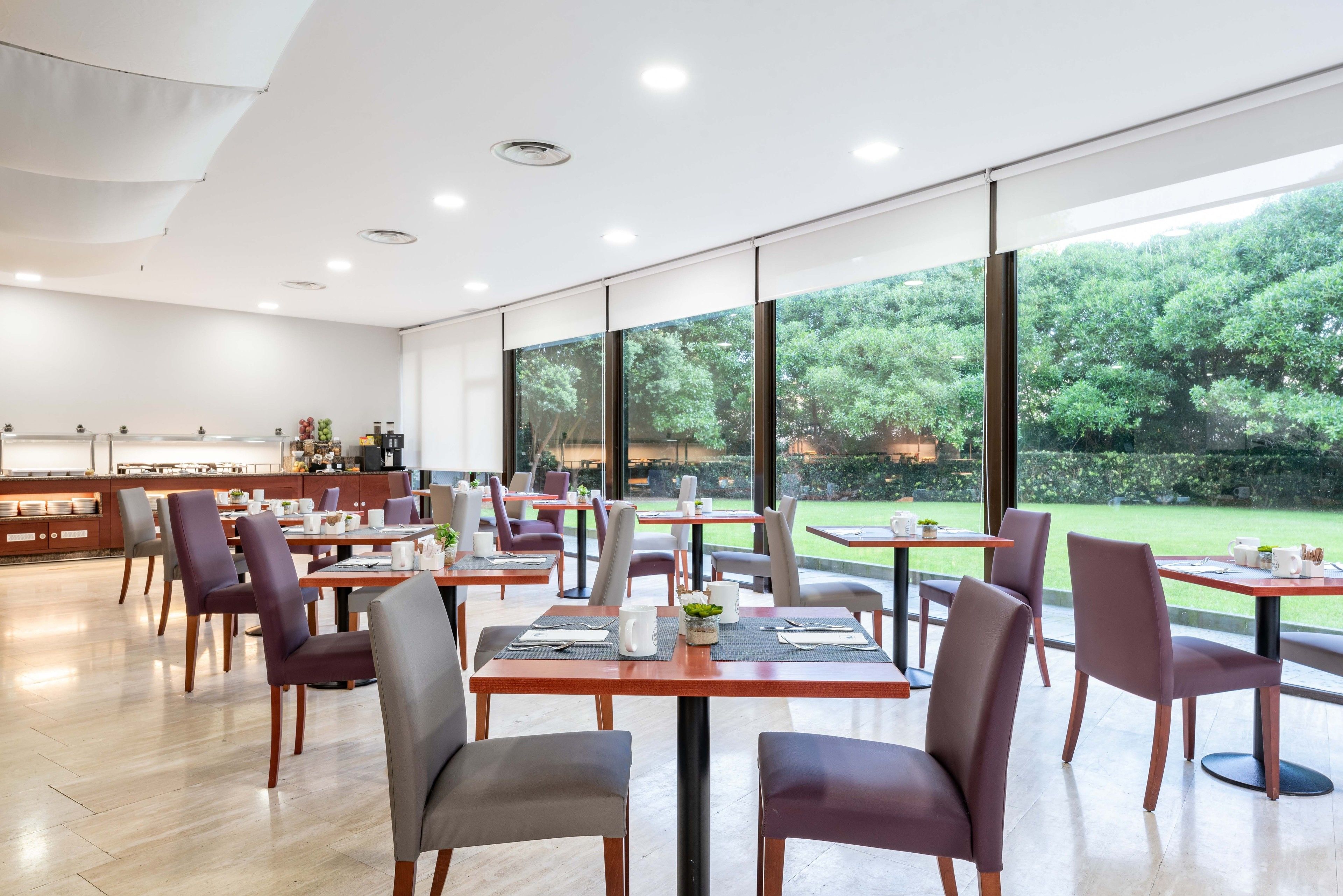 Un comedor moderno con mesas y sillas, ventanas amplias y vistas a zonas verdes exteriores.