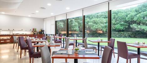 A modern dining area with tables and chairs, large windows, and a view of greenery outside.
