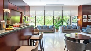 Rebedor modern d'hotel amb zona de restauració, seients i grans finestres que donen vista a vegetació.