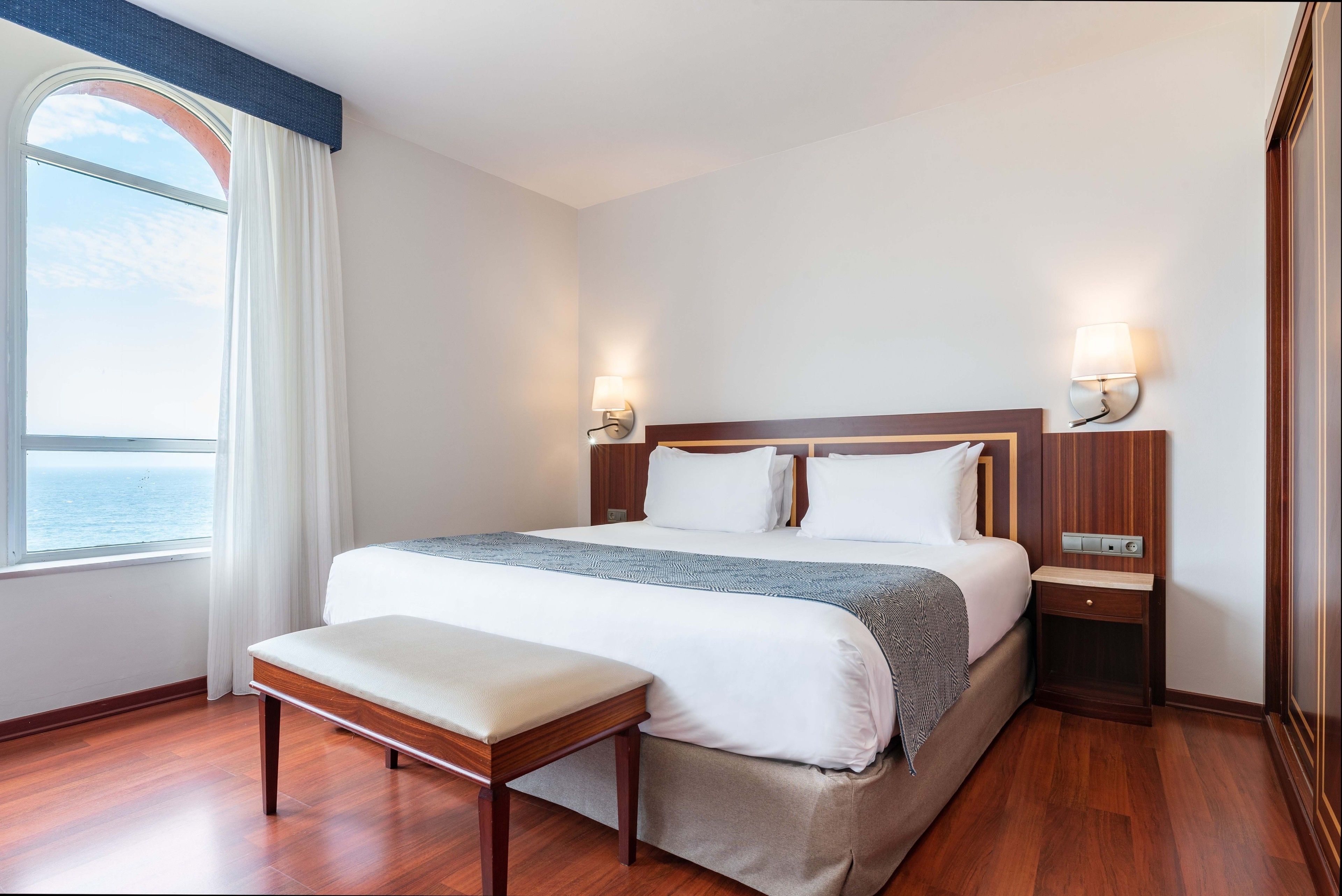 Habitación de hotel con una cama grande, un banco, dos mesitas de noche y vistas al mar.