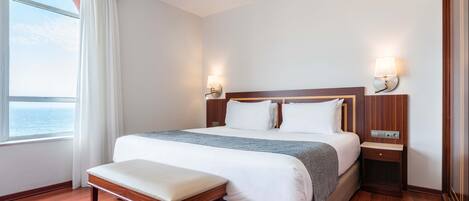 Habitación de hotel con una cama grande, un banco, dos mesitas de noche y vistas al mar.