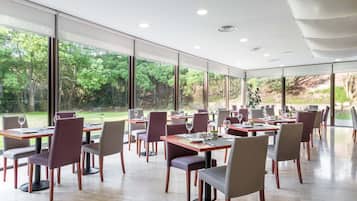 Un comedor moderno con mesas y sillas, ventanas amplias y vistas a zonas verdes al exterior.