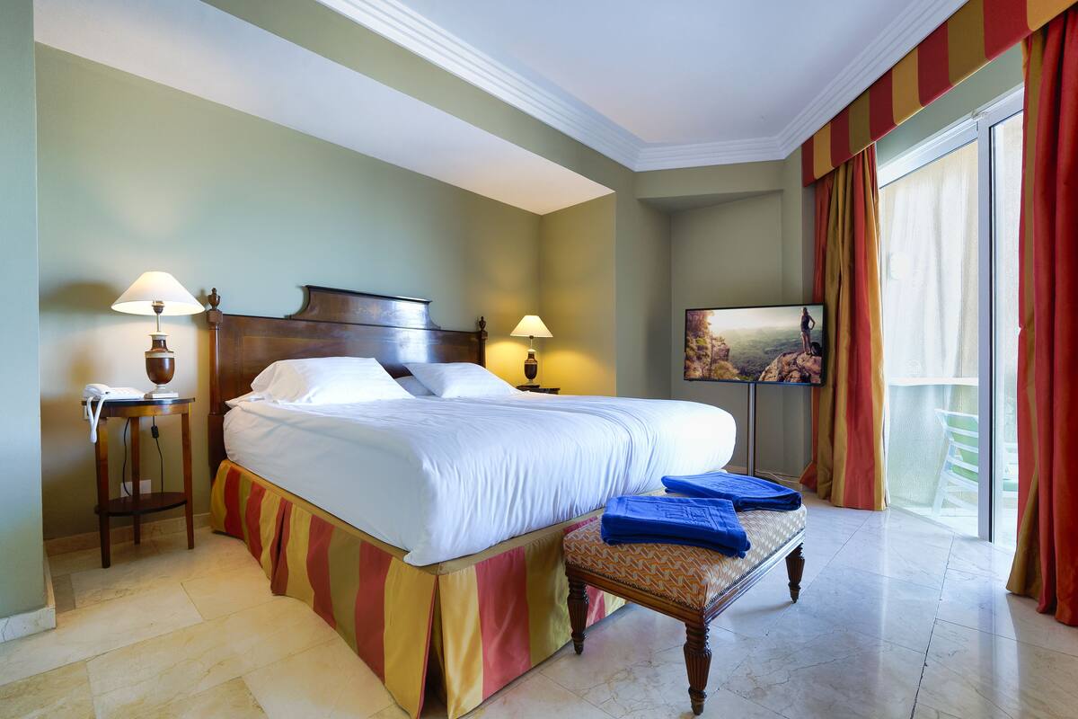 Une chambre d’hôtel avec un grand lit, des lampes de chevet, une petite table sur laquelle est posée une serviette bleue, et un tableau accroché au mur.