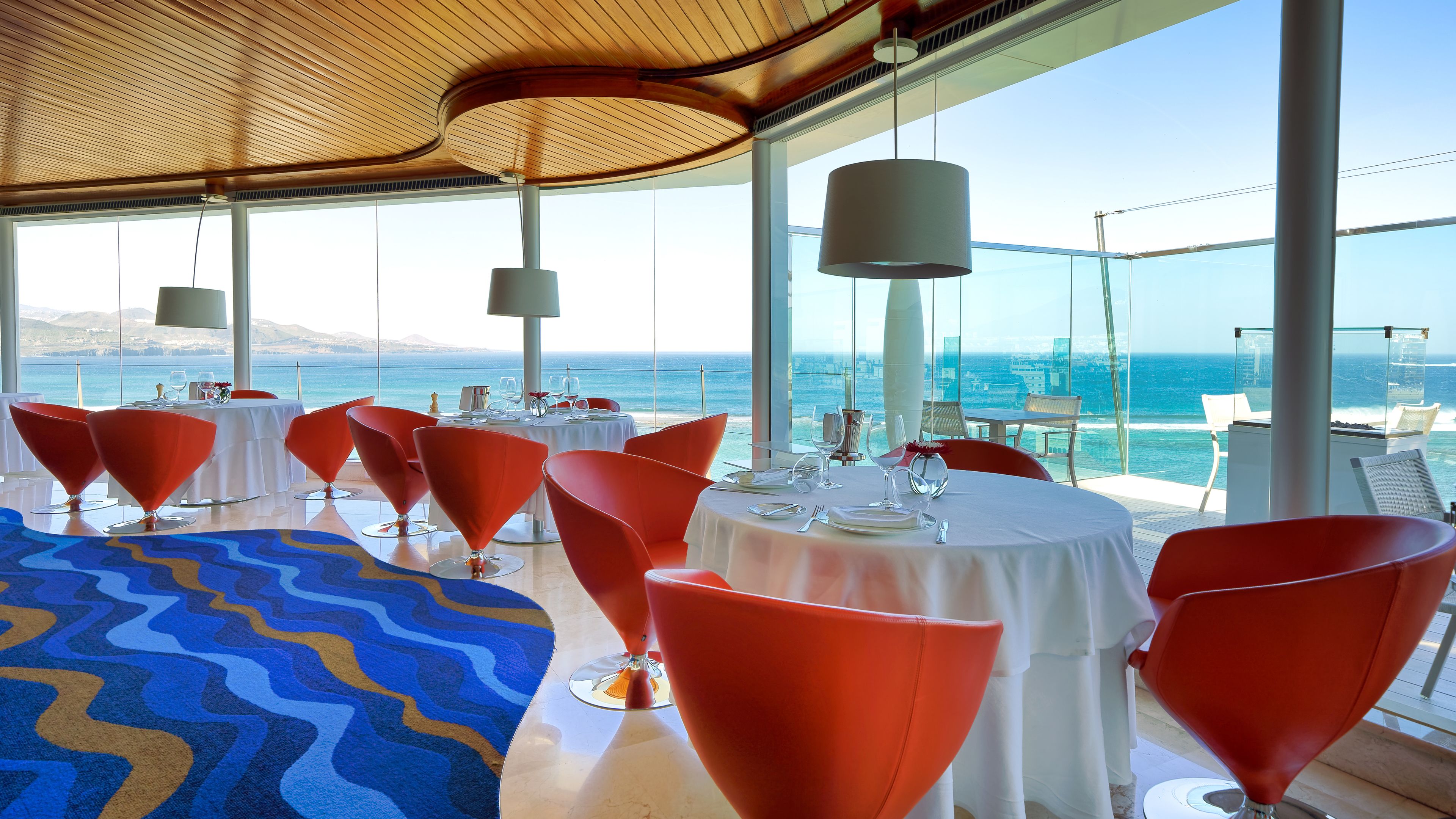 Un restaurante con mesas redondas y sillas rojas, amplias ventanas con vistas al mar y una alfombra azul estampada.