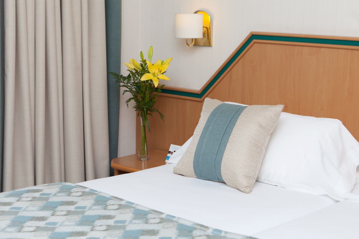 A hotel room with a neatly made bed, a vase with flowers, and a nightstand with a lamp.