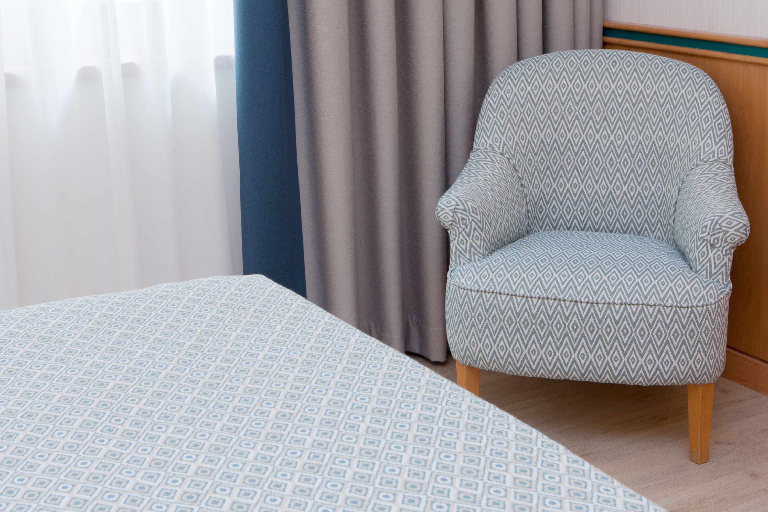 Una habitación con una cama, un sillón estampado, una cortina y un cabecero de madera.