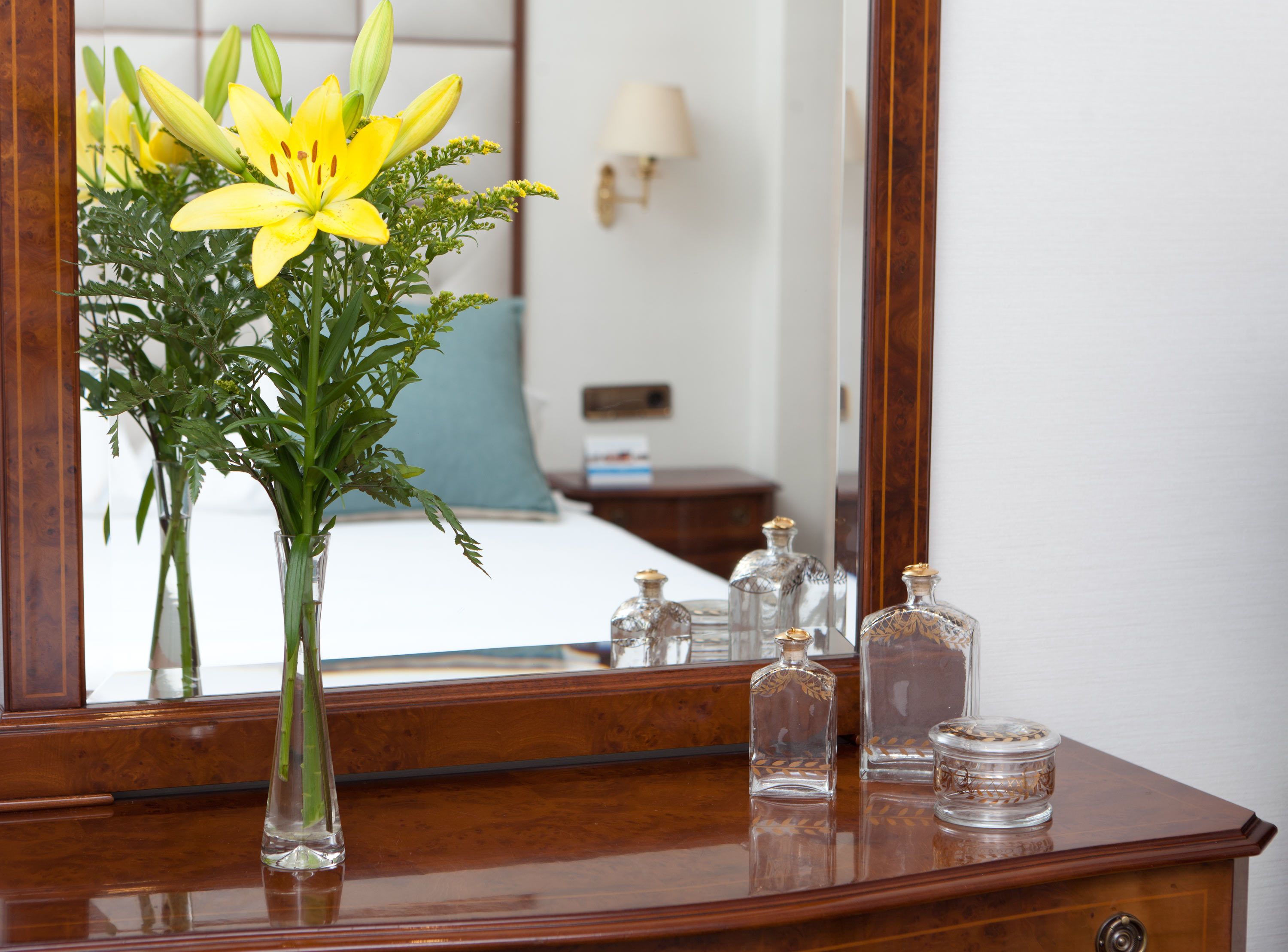Ein hölzerner Kommode mit einem Blumenstrauß aus gelben Blumen, ein Spiegel, der den Raum widerspiegelt, und drei Glasflaschen mit Deckeln.