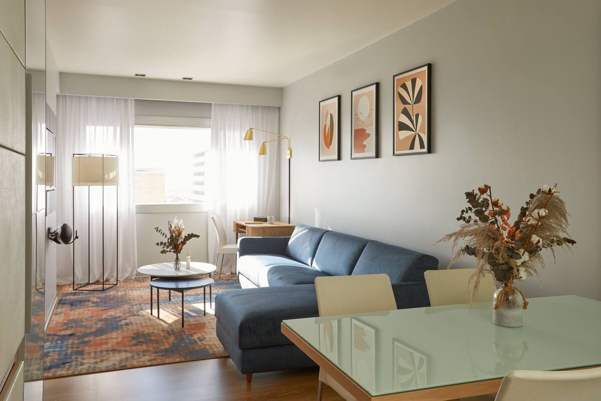 Ein modernes Wohnzimmer mit einem blauen Sofa, einem Glastisch und einer Vase mit Trockenblumen. Ein kleiner runder Tisch mit Lampe und Stuhl steht dort, und ein großes Fenster mit transparenten Vorhängen.