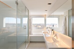 Un baño moderno con cabina de ducha de vidrio, un amplio ventanal con vistas a la ciudad y lavamanos dobles con tocador.