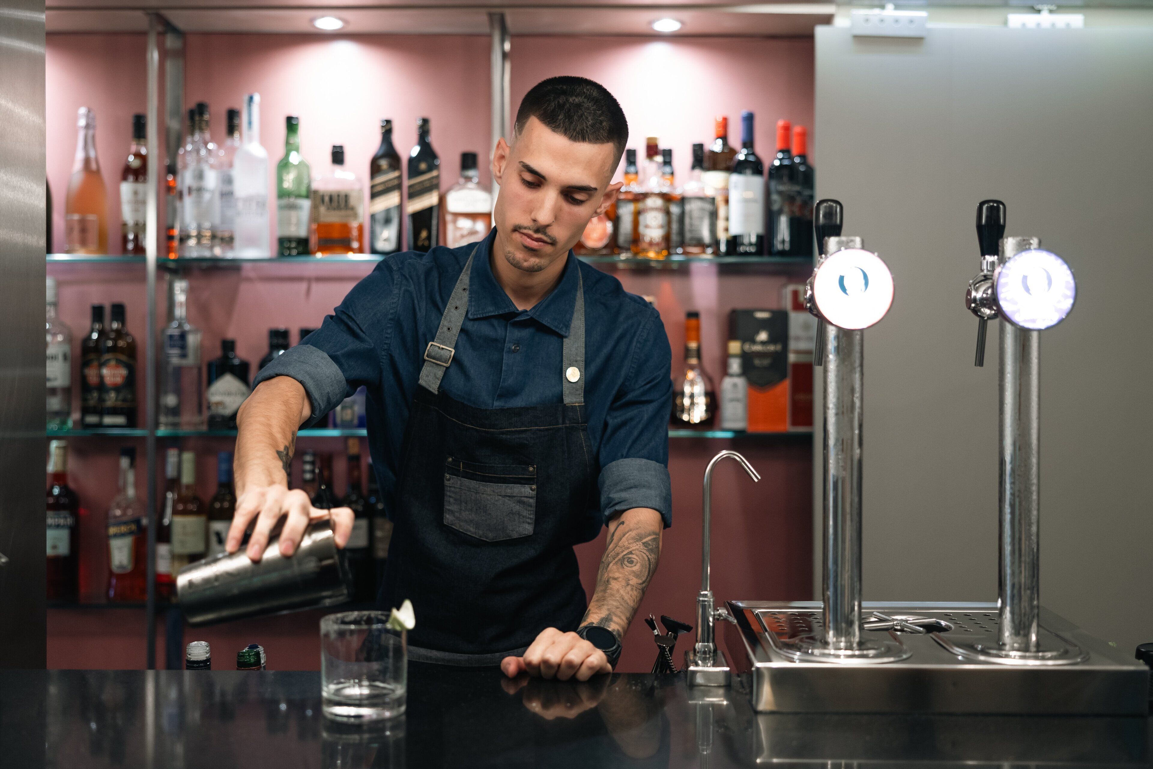 Een barman maakt een drankje klaar achter een bar, met flessen drank op de achtergrond.