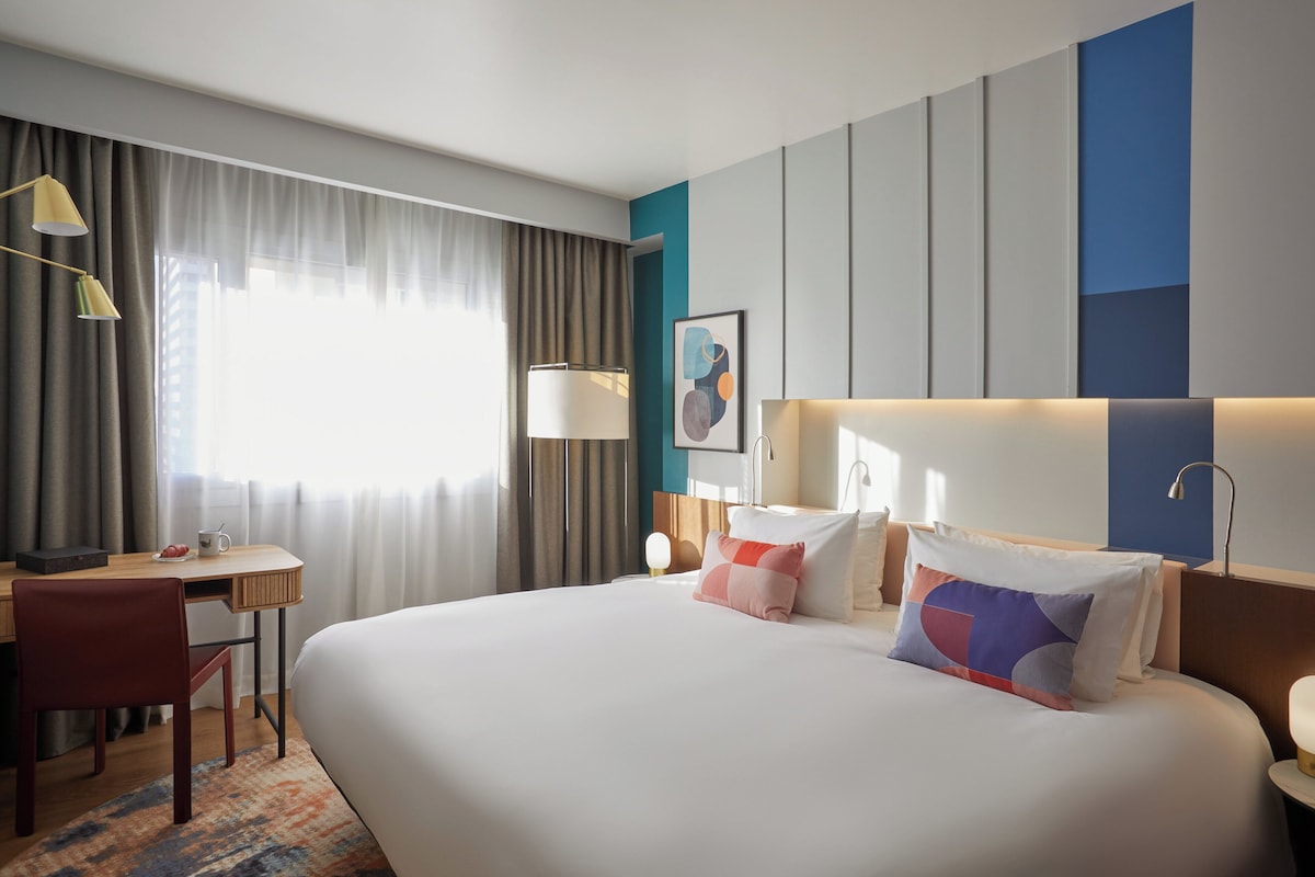 Una camera d'albergo con un letto grande, una scrivania con una sedia, una lampada e un quadro appeso al muro.