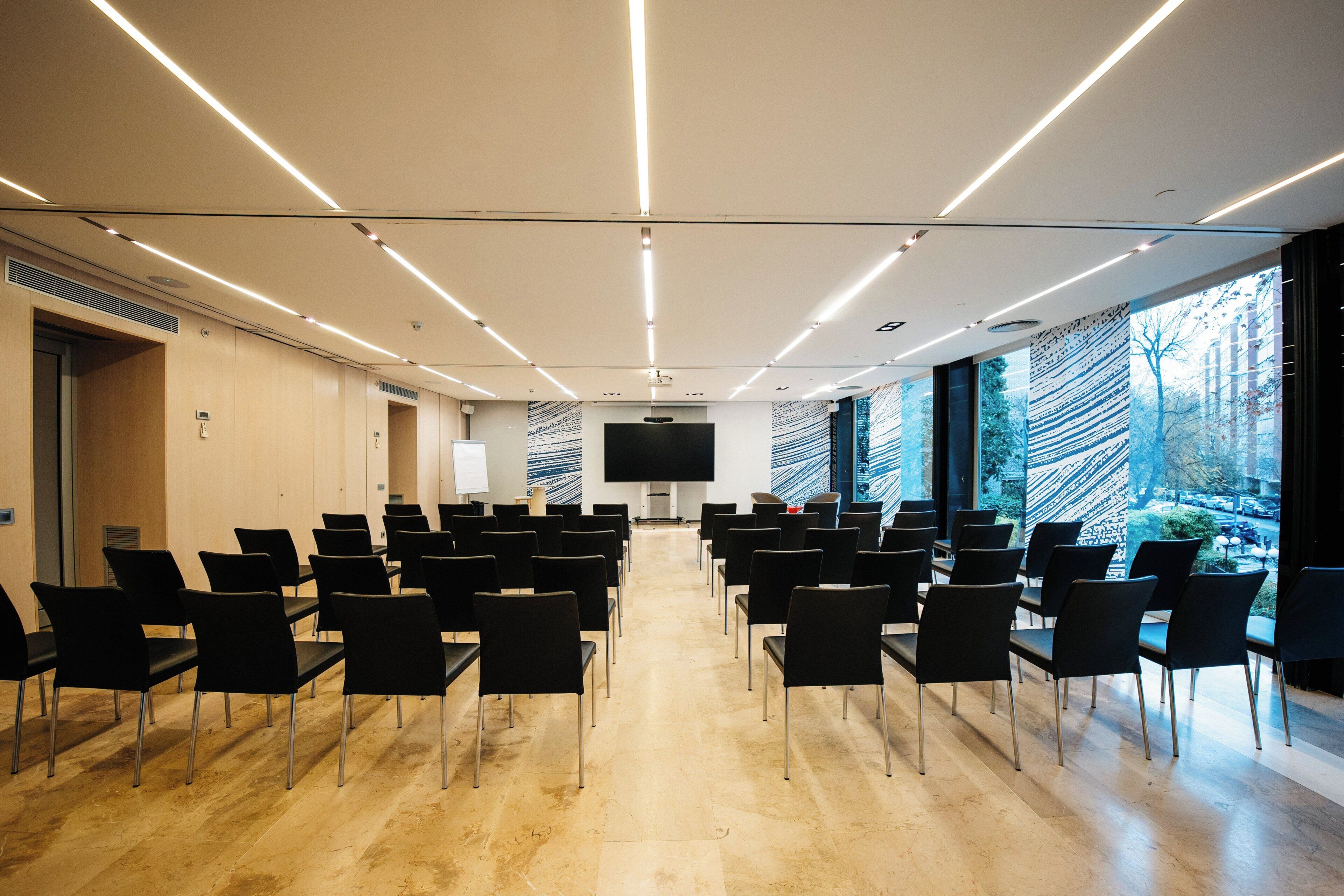 Un salón de conferencias con filas de sillas, una pantalla grande y vistas al exterior.