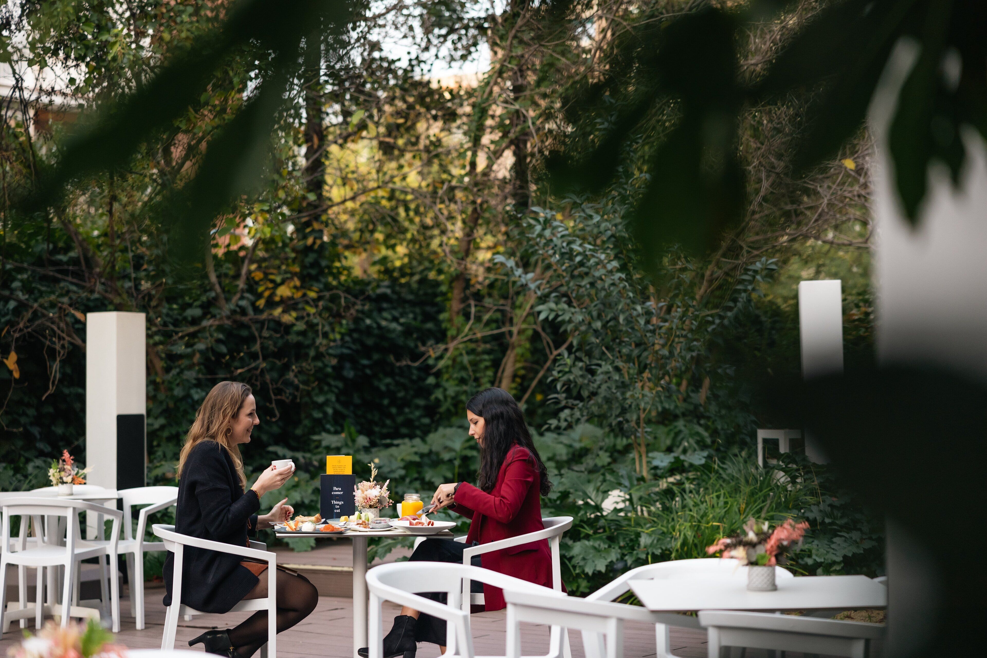Two people dining at an outdoor table with white chairs, surrounded by greenery.