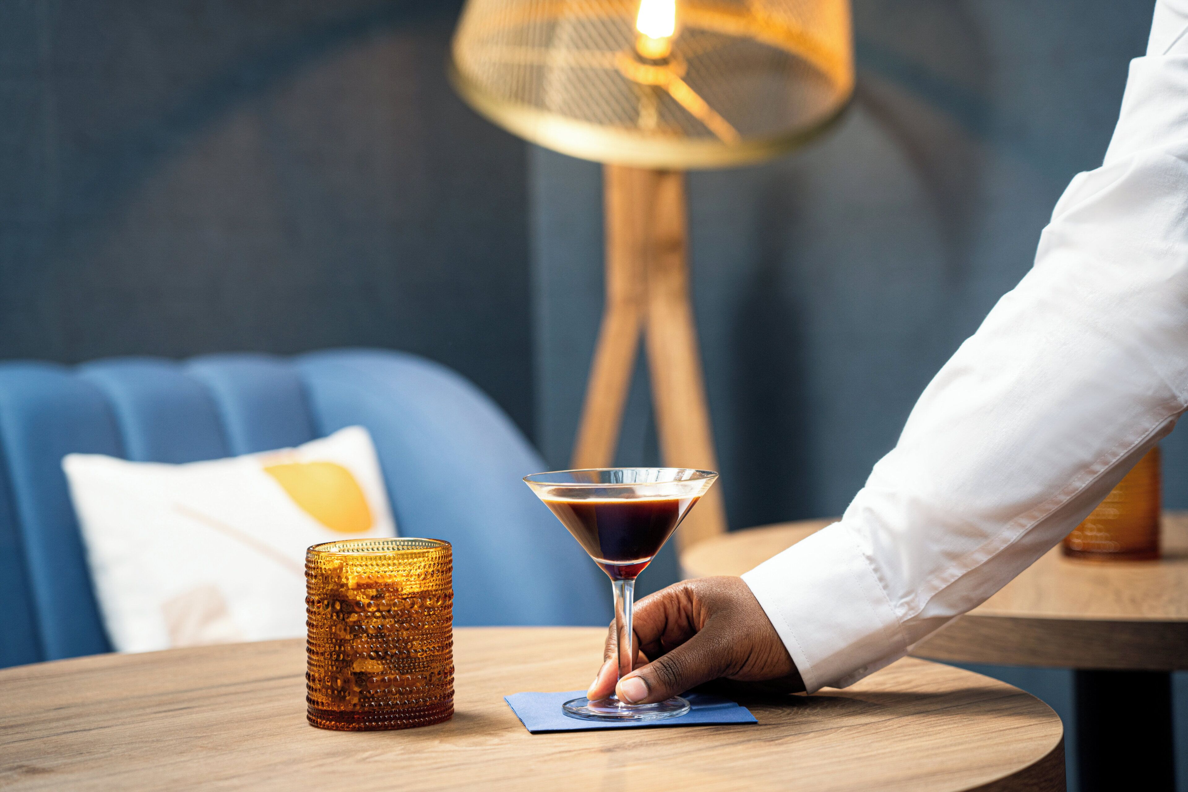Une main tenant un verre à martini sur une table en bois, avec un porte-bougie texturé à côté.