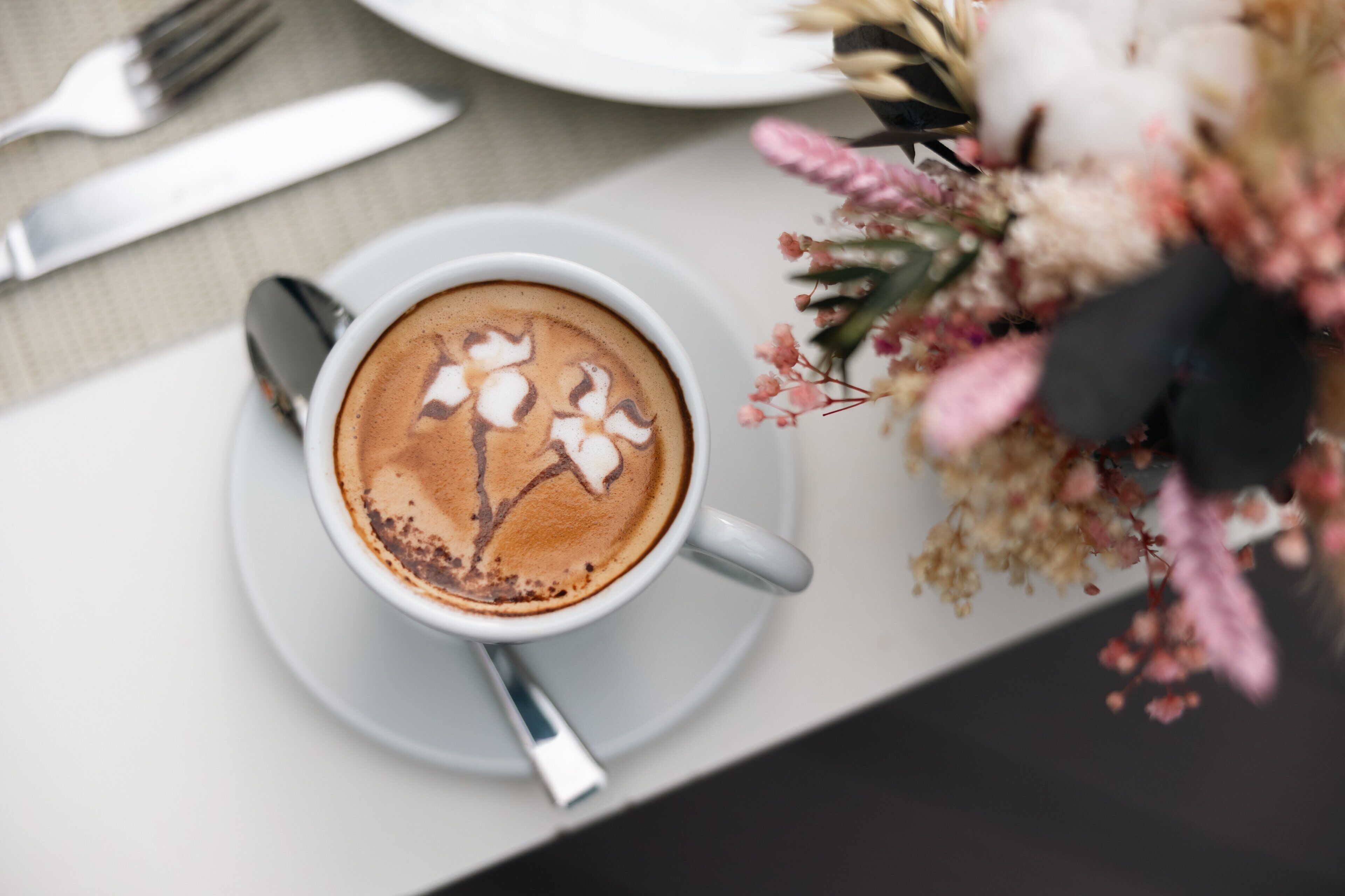 Una tazza di caffè con decorazione in schiuma latte su un piattino, un cucchiaino, una forchetta e un bouquet di fiori su un tavolo.