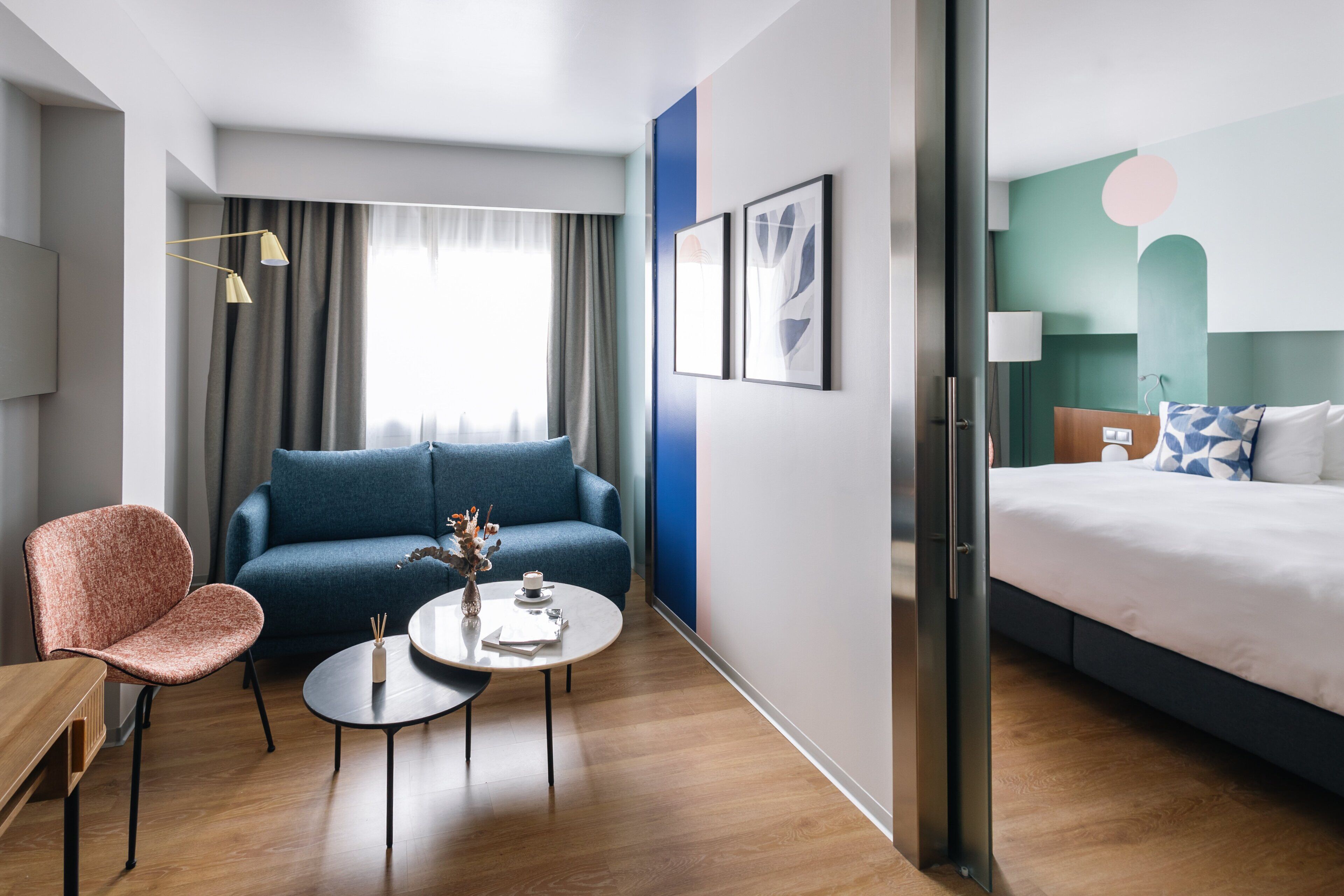 A modern hotel room with a blue sofa, a wooden chair, a small round table, and a bed with white bedding.