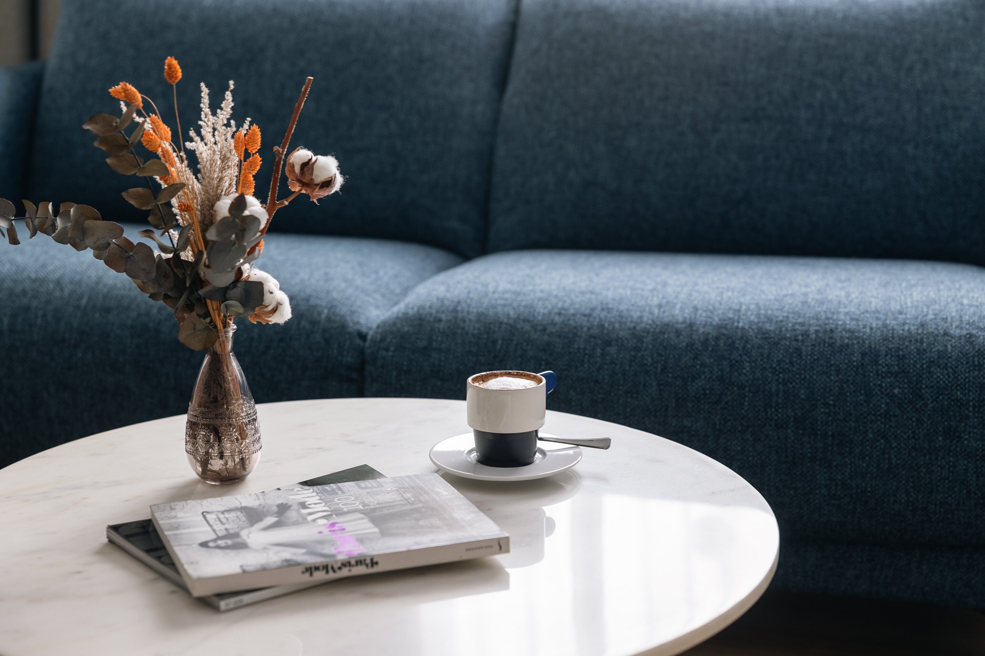 Una mesa redonda blanca con una taza de café, un libro y un jarrón con flores secas.