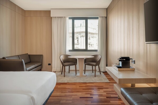 A hotel room with a bed, a sofa, a dining table with chairs, a coffee machine, and a TV mounted on the wall.