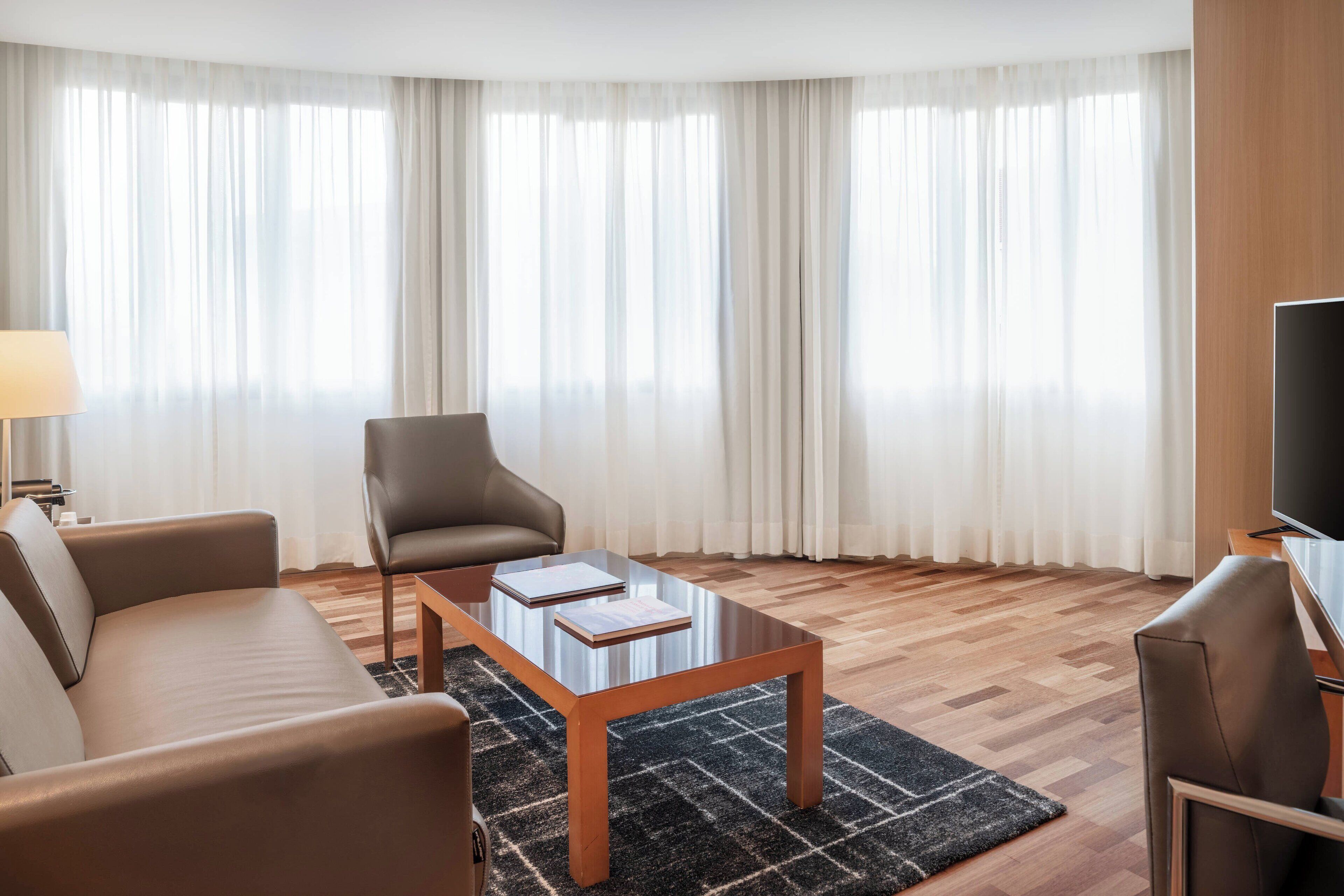 Una sala de estar moderna con un sofá, un sillón y una mesa de centro, un televisor de pantalla plana y ventanas amplias con cortinas translúcidas.