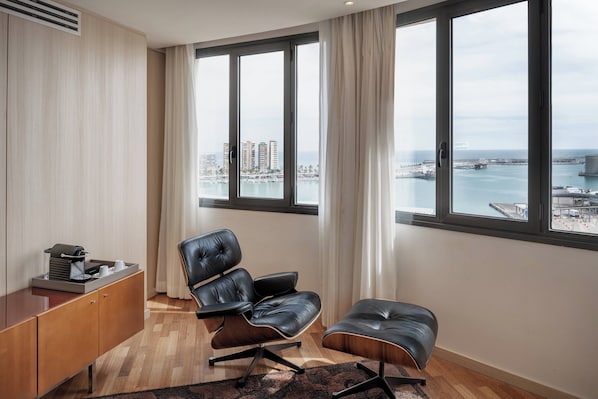 Una habitación moderna con un ventanal grande, un sillón de cuero, un pequeño reposapiés y un armario de madera.