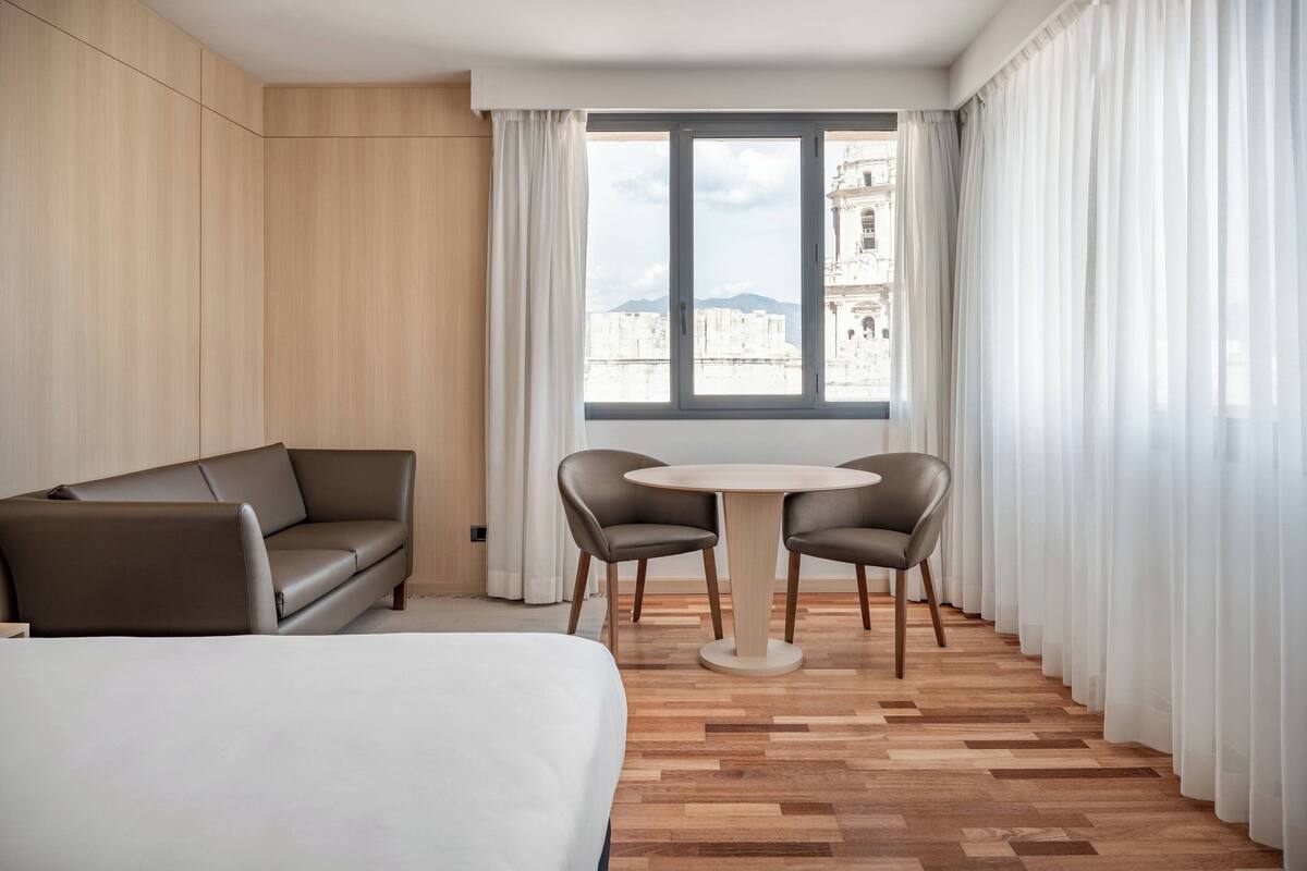 Hotelkamer met een bed, een bank, een ronde tafel met stoelen en uitzicht op een gebouw door een groot raam.