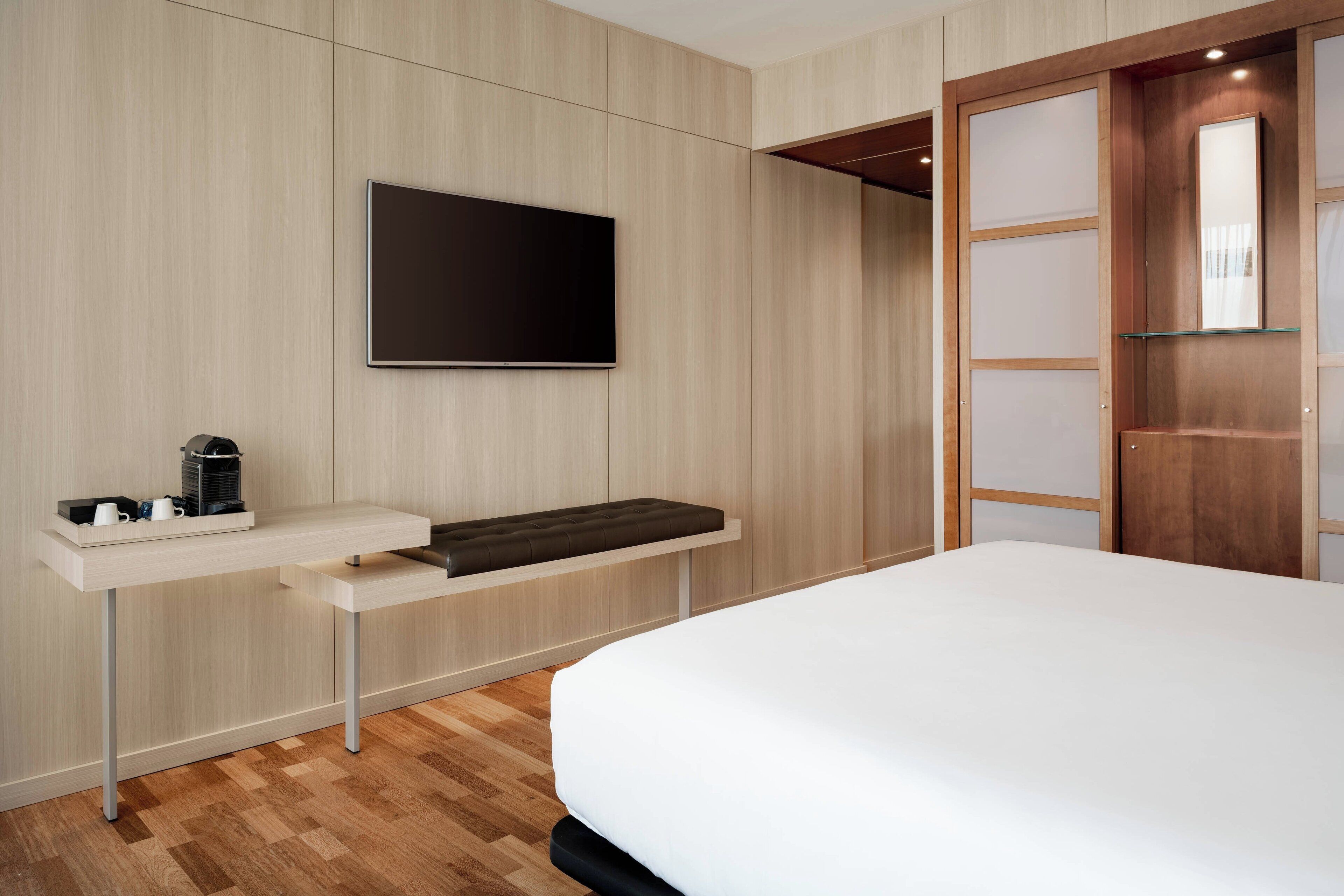 A modern hotel room with a bed, a flat-screen TV mounted on the wall, a small table with a coffee maker, and a wooden wardrobe with sliding doors.