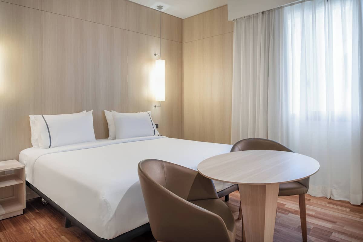 Hotelkamer met groot bed, twee kussens, nachtkastje, stoel, ronde tafel en raam met gordijnen.