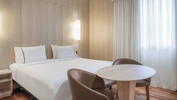 A hotel room with a large bed, two pillows, a bedside table, a chair, a round table, and a window with curtains.