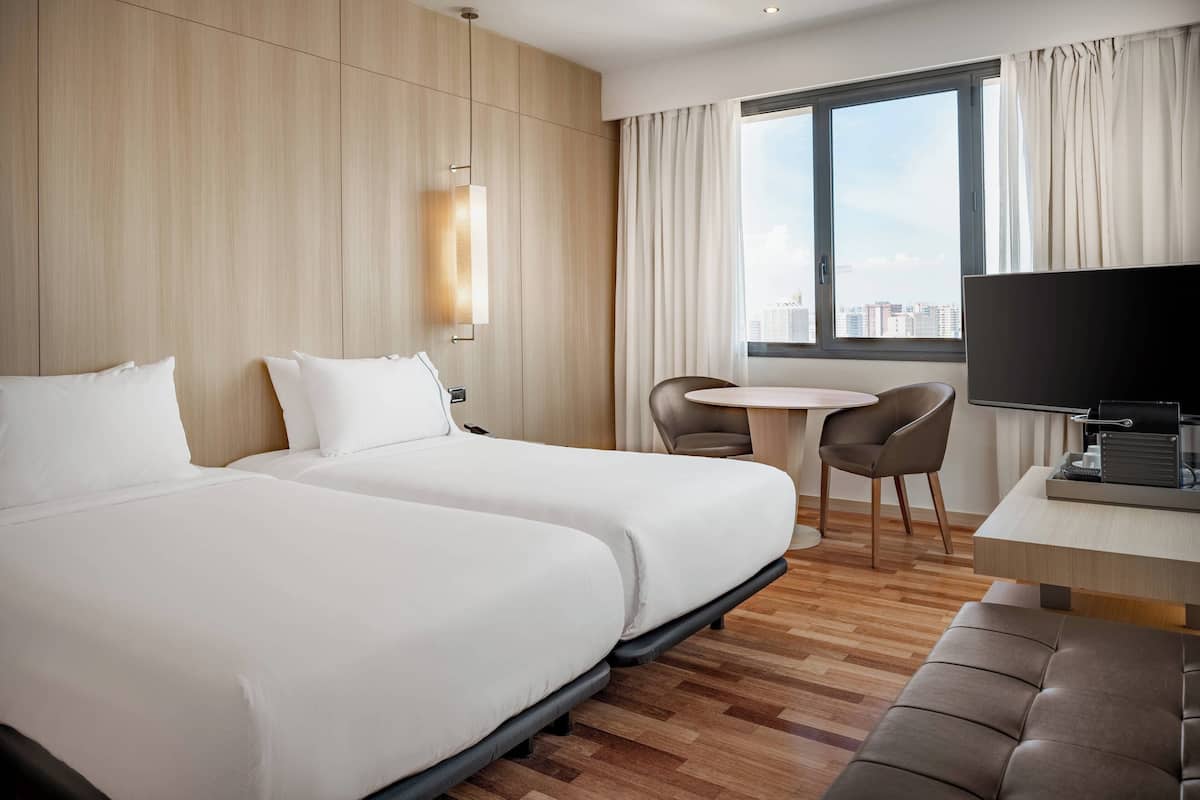 Hotelkamer met twee bedden, een bureau, een stoel en een groot raam met uitzicht op de stad.