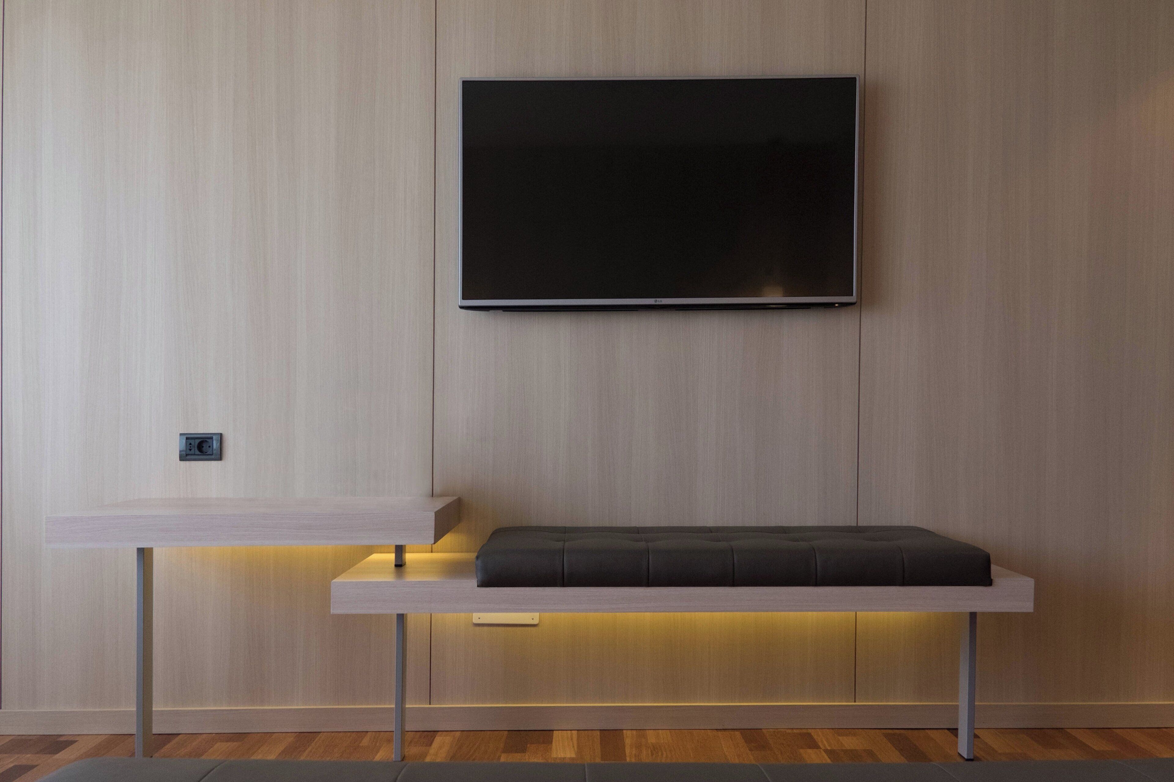Une chambre moderne dotée d’un téléviseur à écran plat fixé au mur, d’un banc en bois avec un coussin et d’une étagère intégrée en dessous.