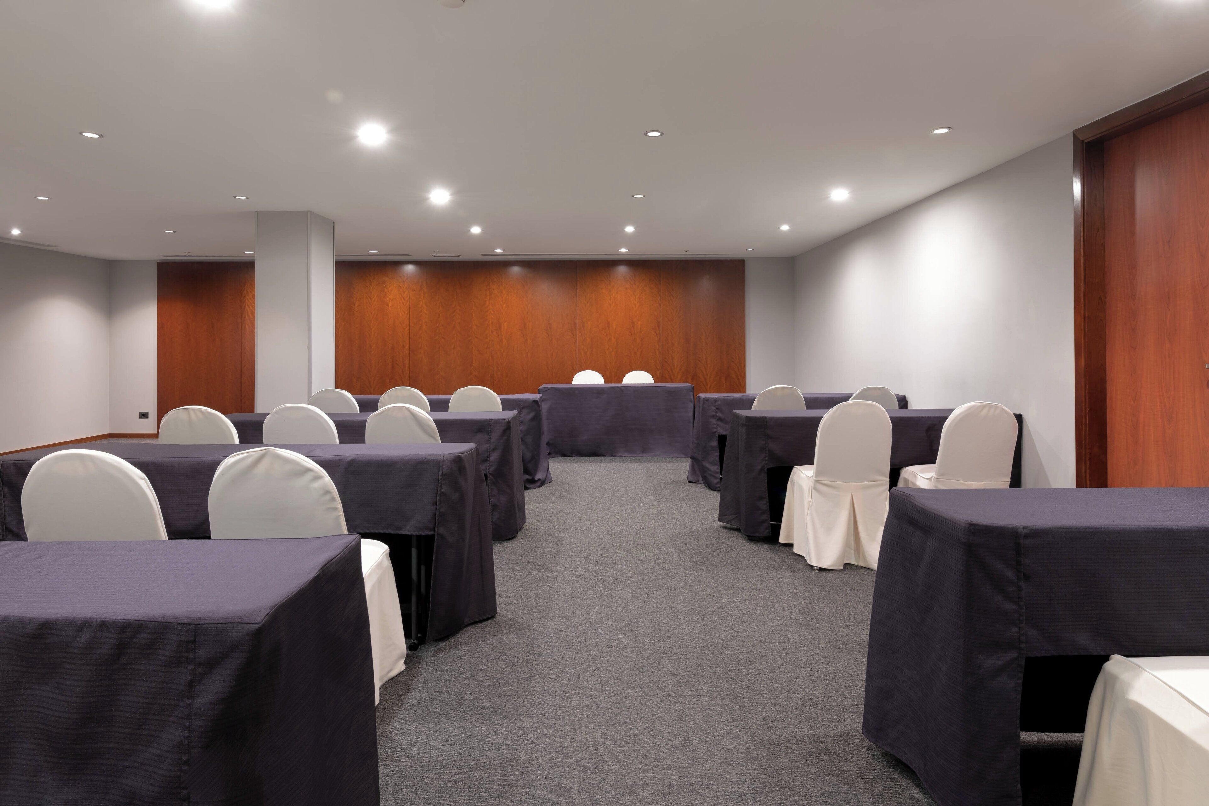Un salón de conferencias con filas de mesas y sillas, piso alfombrado, paneles de madera y luces empotradas en el techo.