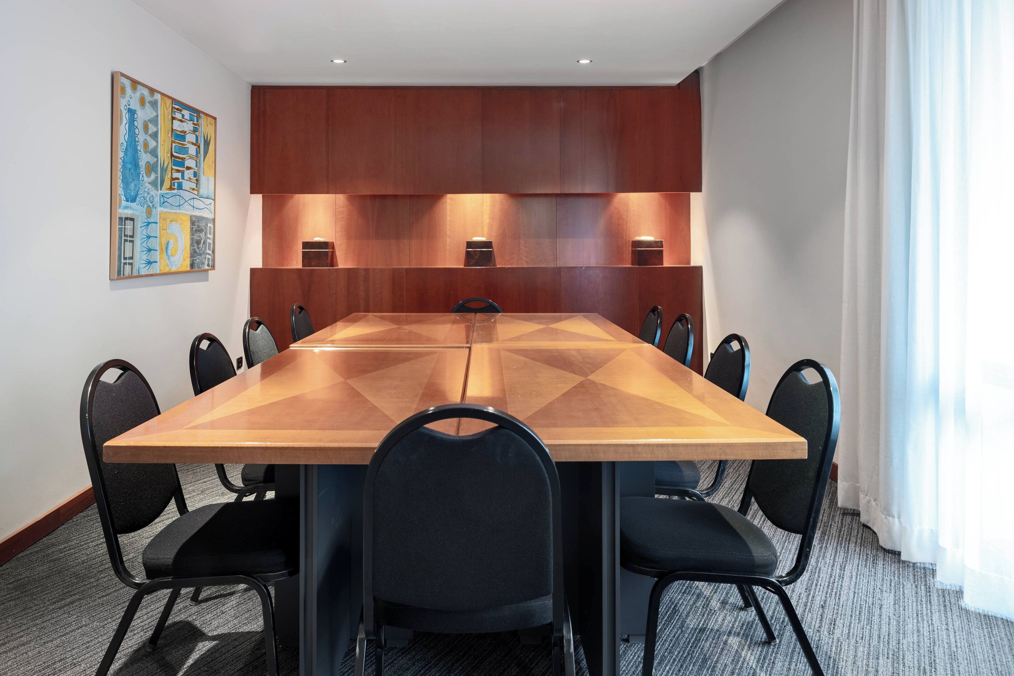A conference room with a large wooden table, black chairs, recessed lighting, and a painting on the wall.