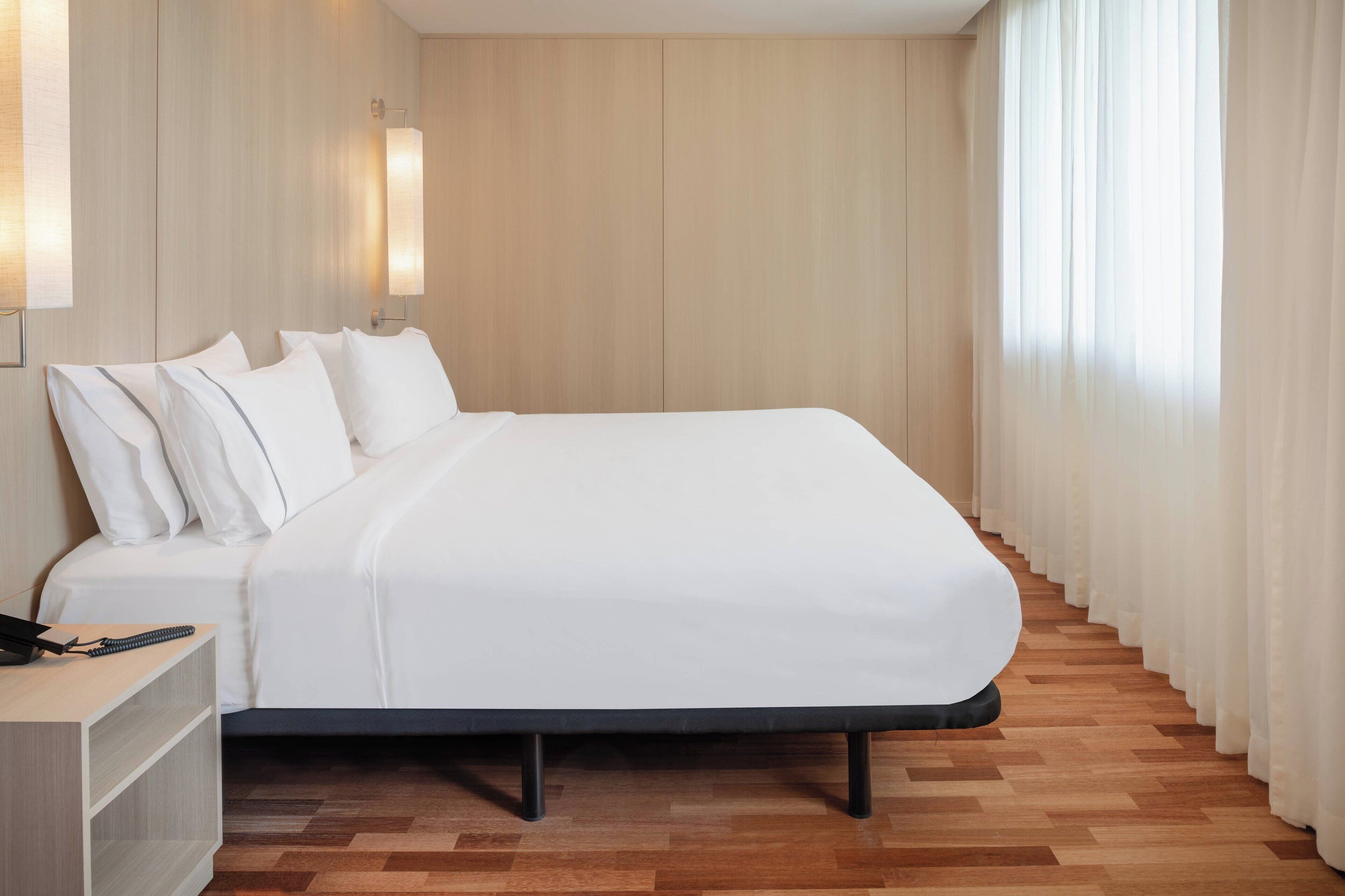 A modern hotel room with a large bed, wooden flooring, and a nightstand with a phone and remote control.
