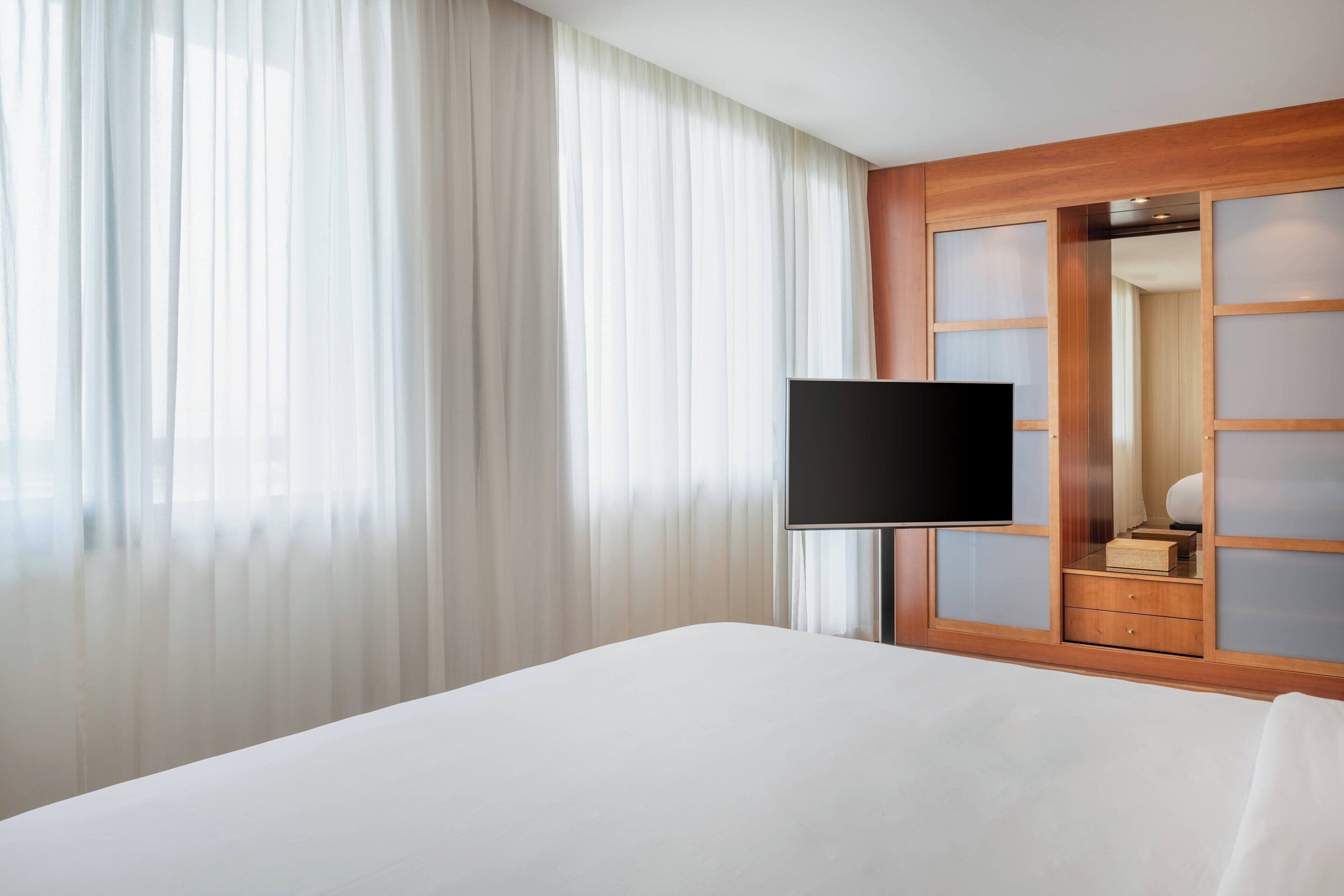 Una habitación de hotel con una cama grande, un televisor de pantalla plana, un armario de madera y cortinas traslúcidas.