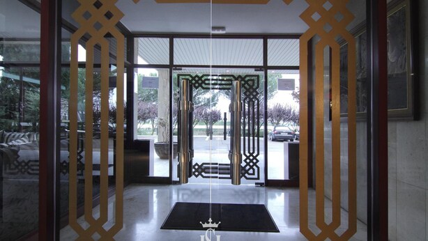Entrada de hotel con puerta de vidrio, felpudo negro con logotipo de una corona y una reja de metal.