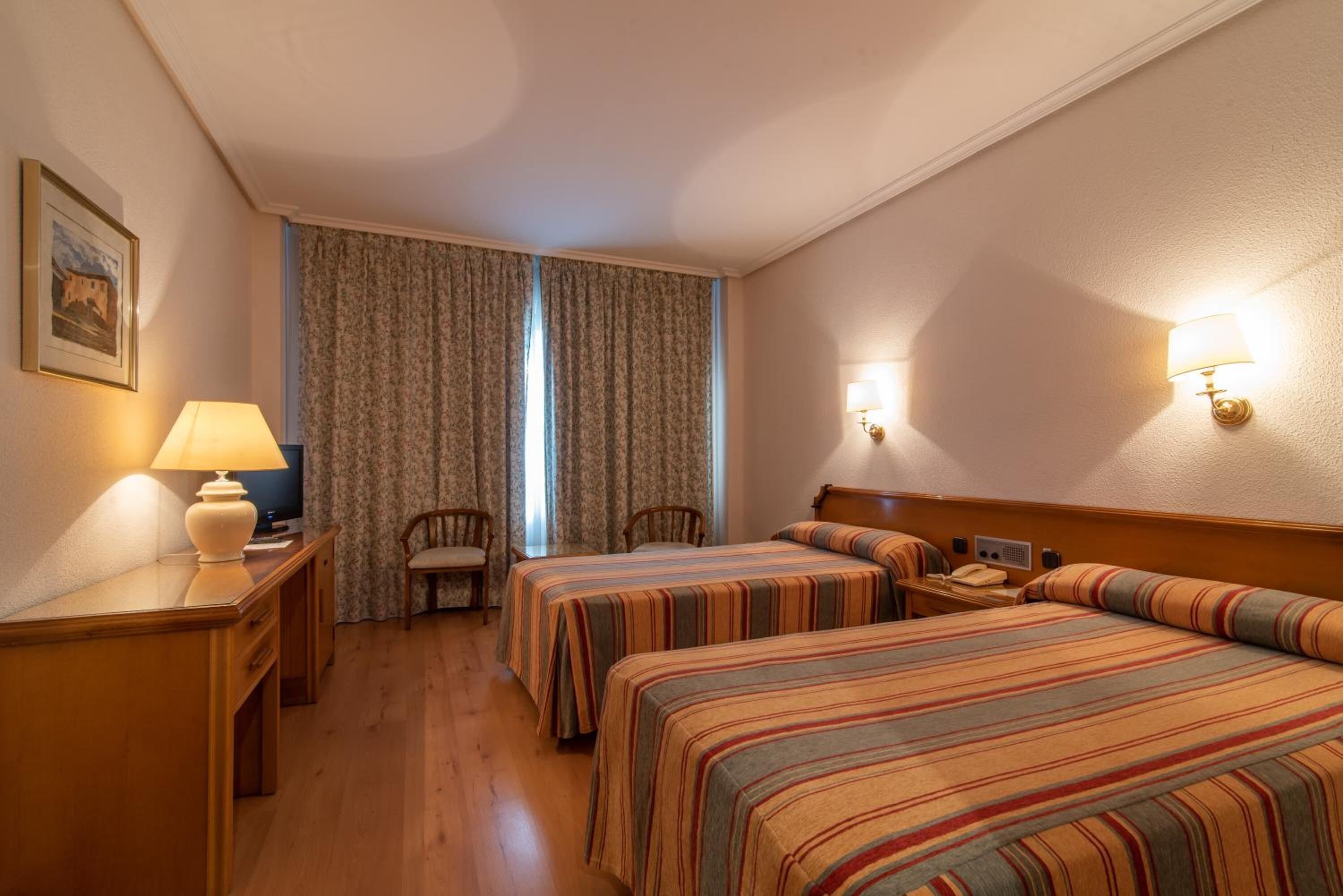 Ein Hotelzimmer mit zwei Betten, einem Schreibtisch, einem Stuhl, einer Lampe und einem Bild an der Wand.