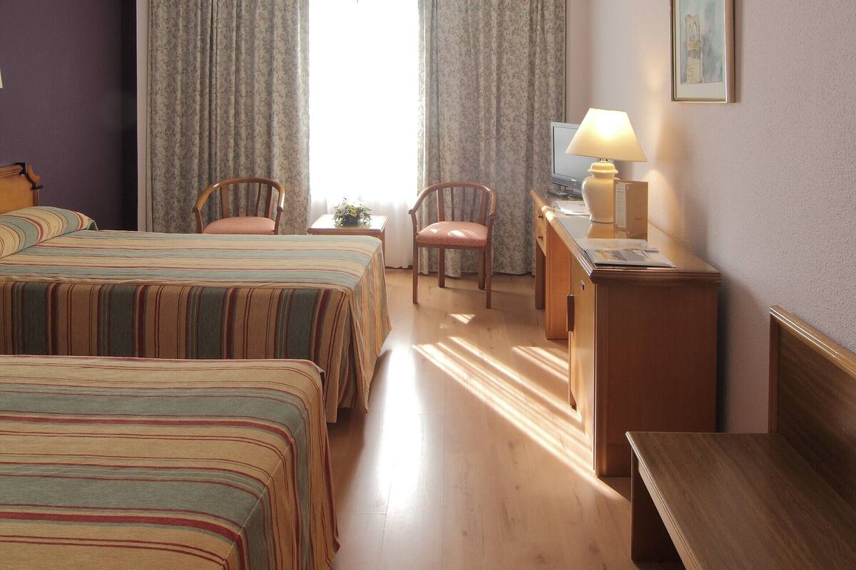 A hotel room with two beds, a desk with a lamp, a chair, and a window with curtains.