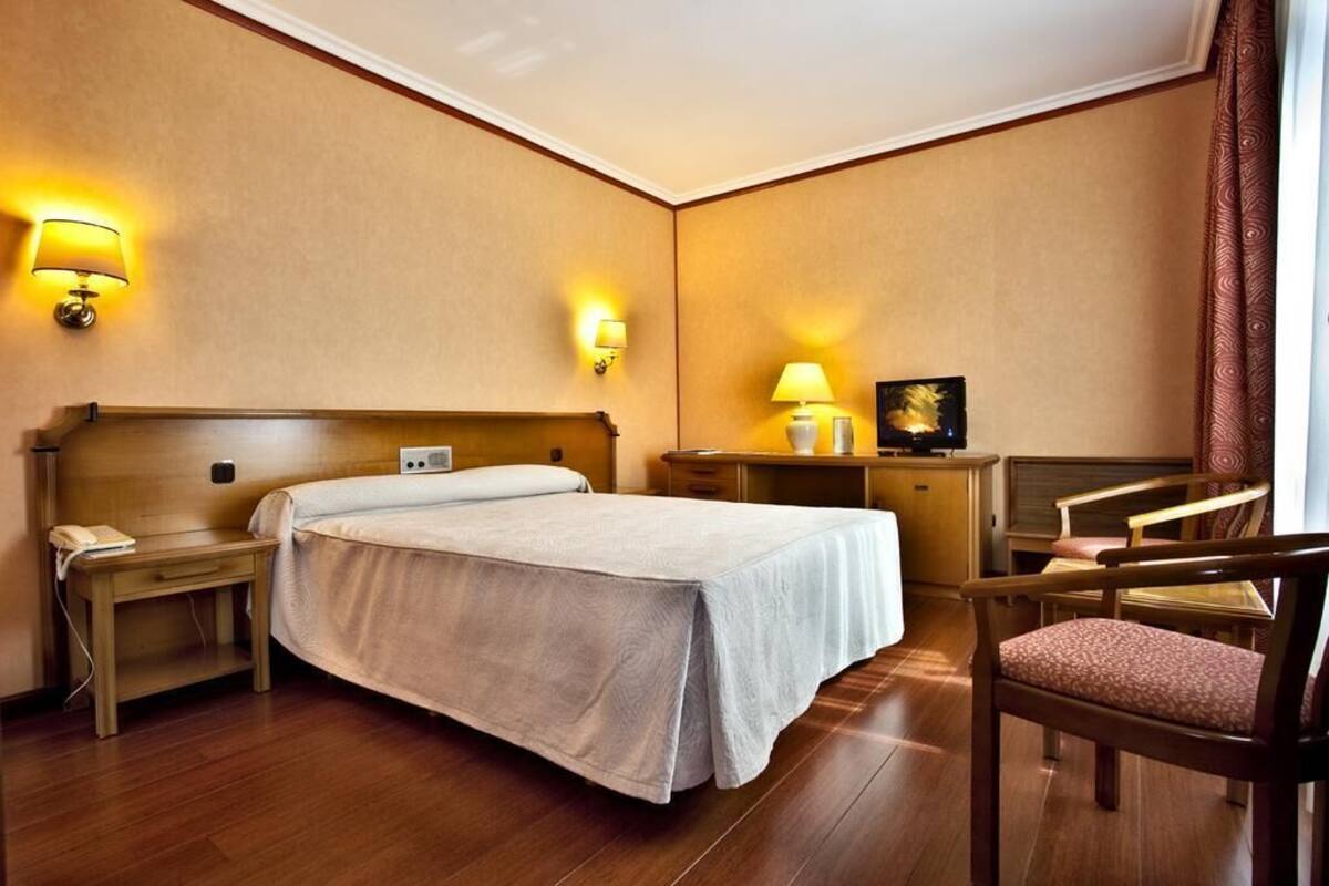 A hotel room with a bed, bedside tables, a desk, a chair, and wall-mounted lamps.