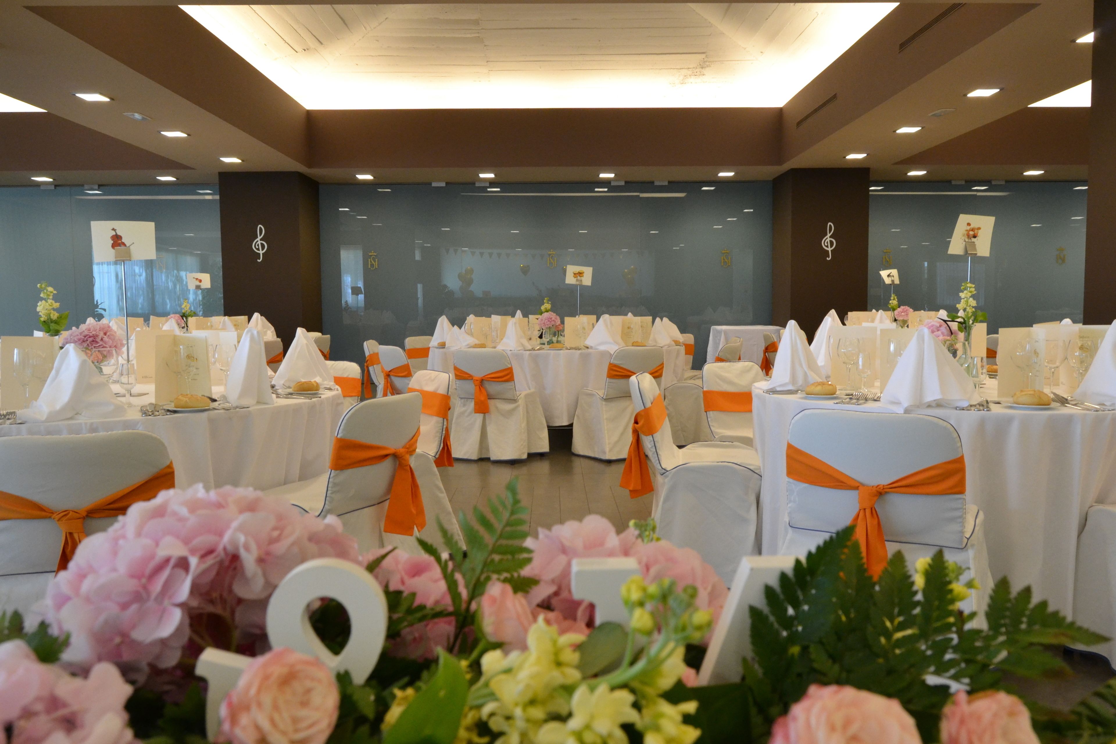Un salón de banquetes con mesas redondas, manteles blancos y cintas anaranjadas.
